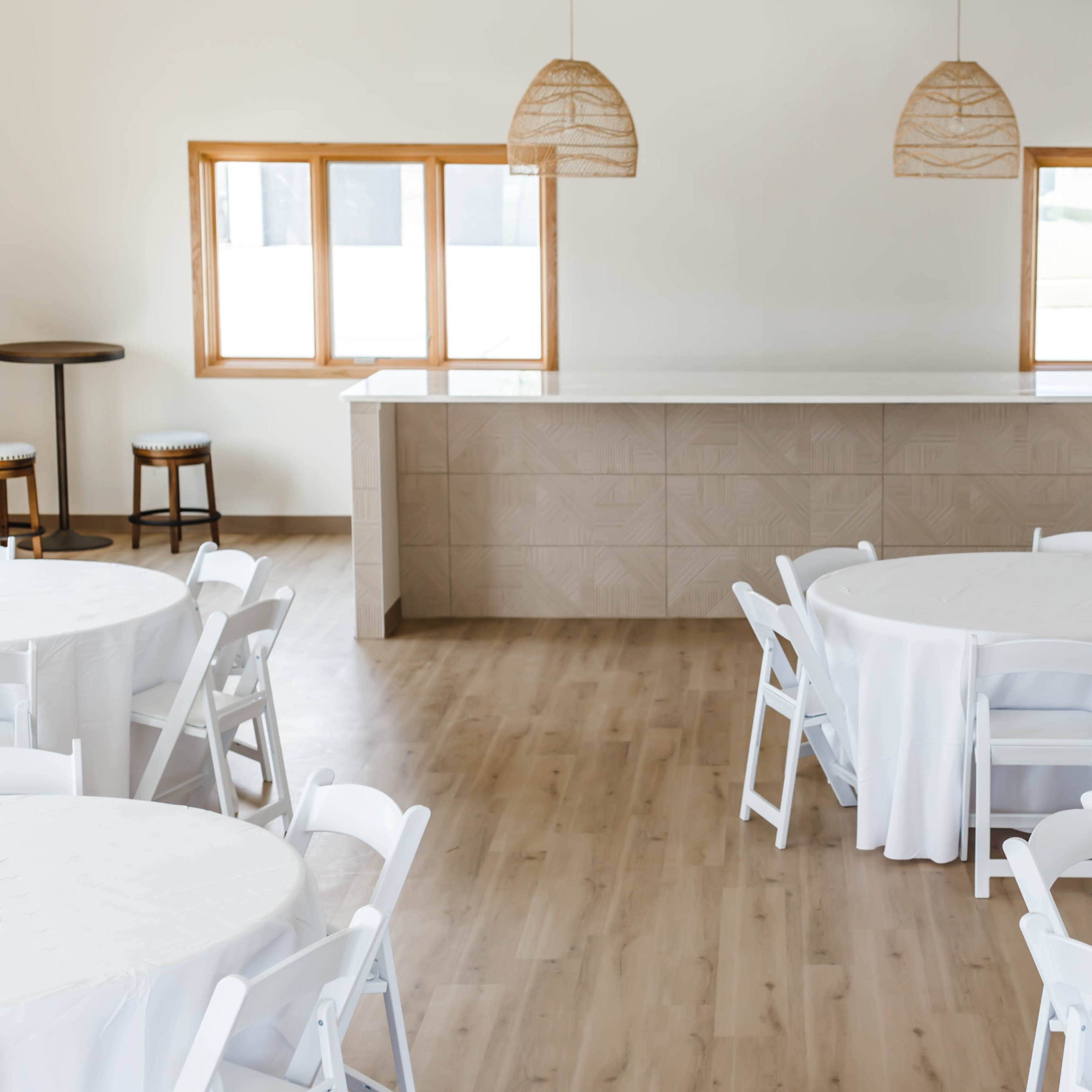 A spacious indoor event venue with round tables covered in white tablecloths, a light-colored bar area in the background, and large windows allowing natural light to enter.