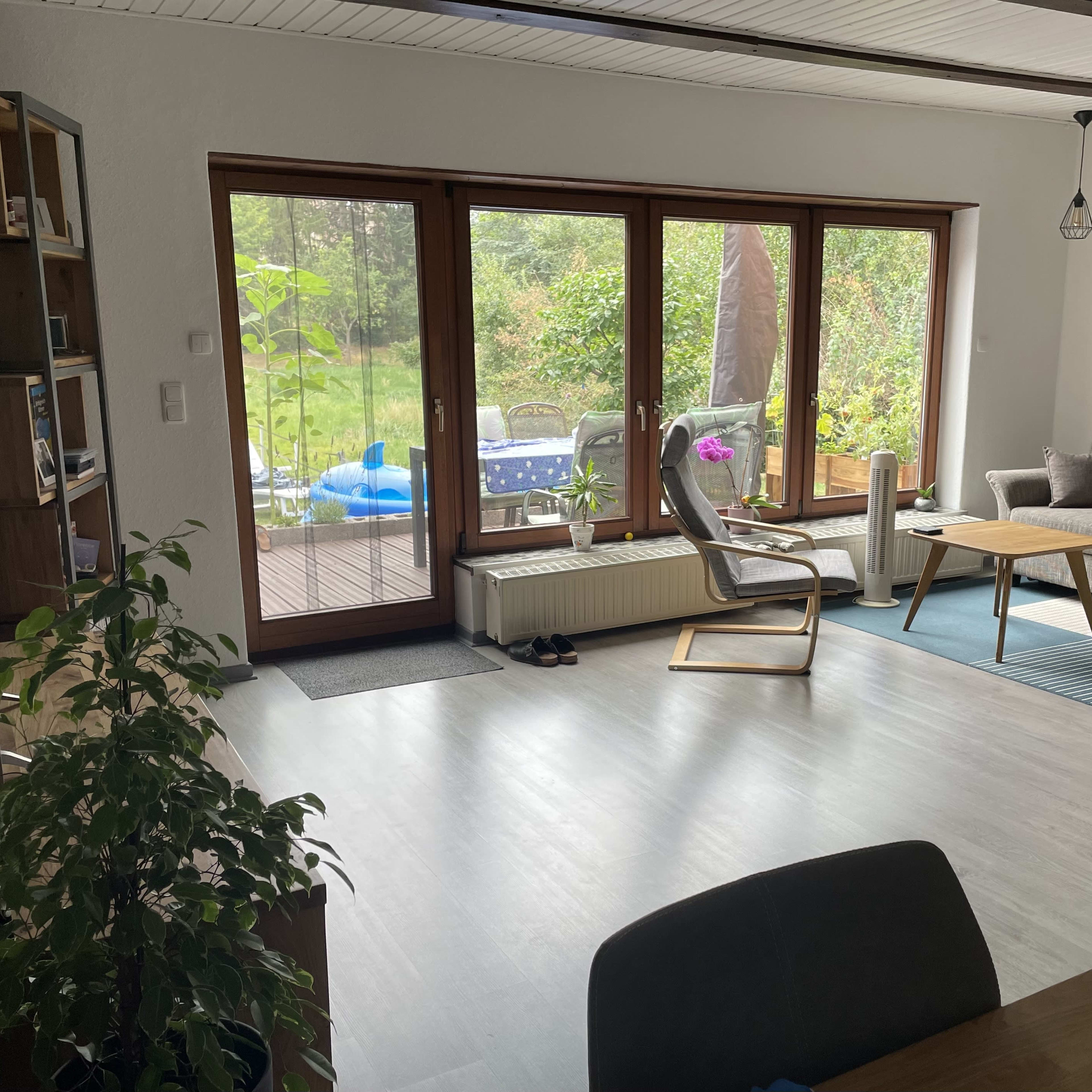 A living room with large windows, a couch, a wooden table, and a view of a garden outside.