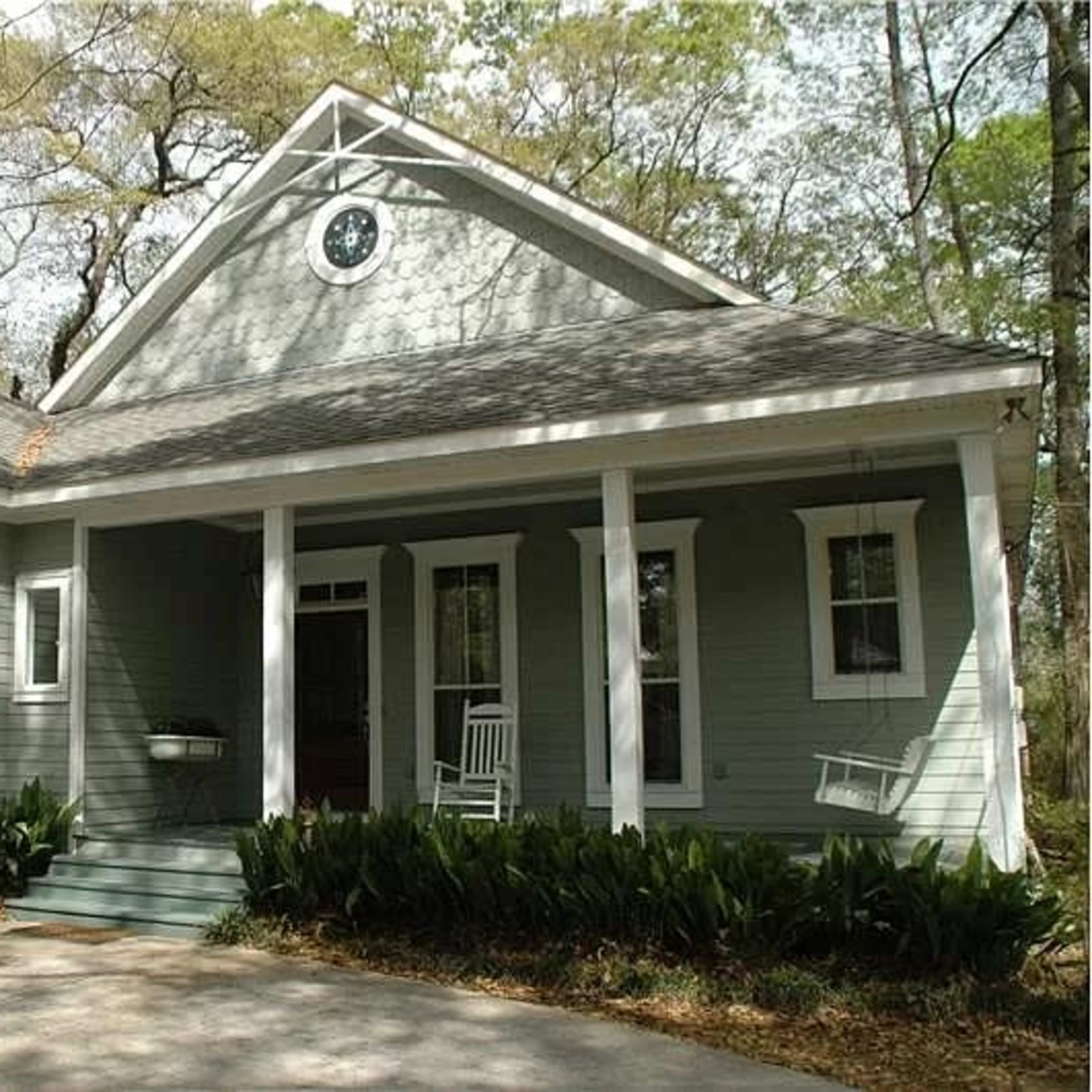 A pale green house with a gabled roof, front porch, and white rocking chairs on either side of the entrance, surrounded by greenery and bare trees.