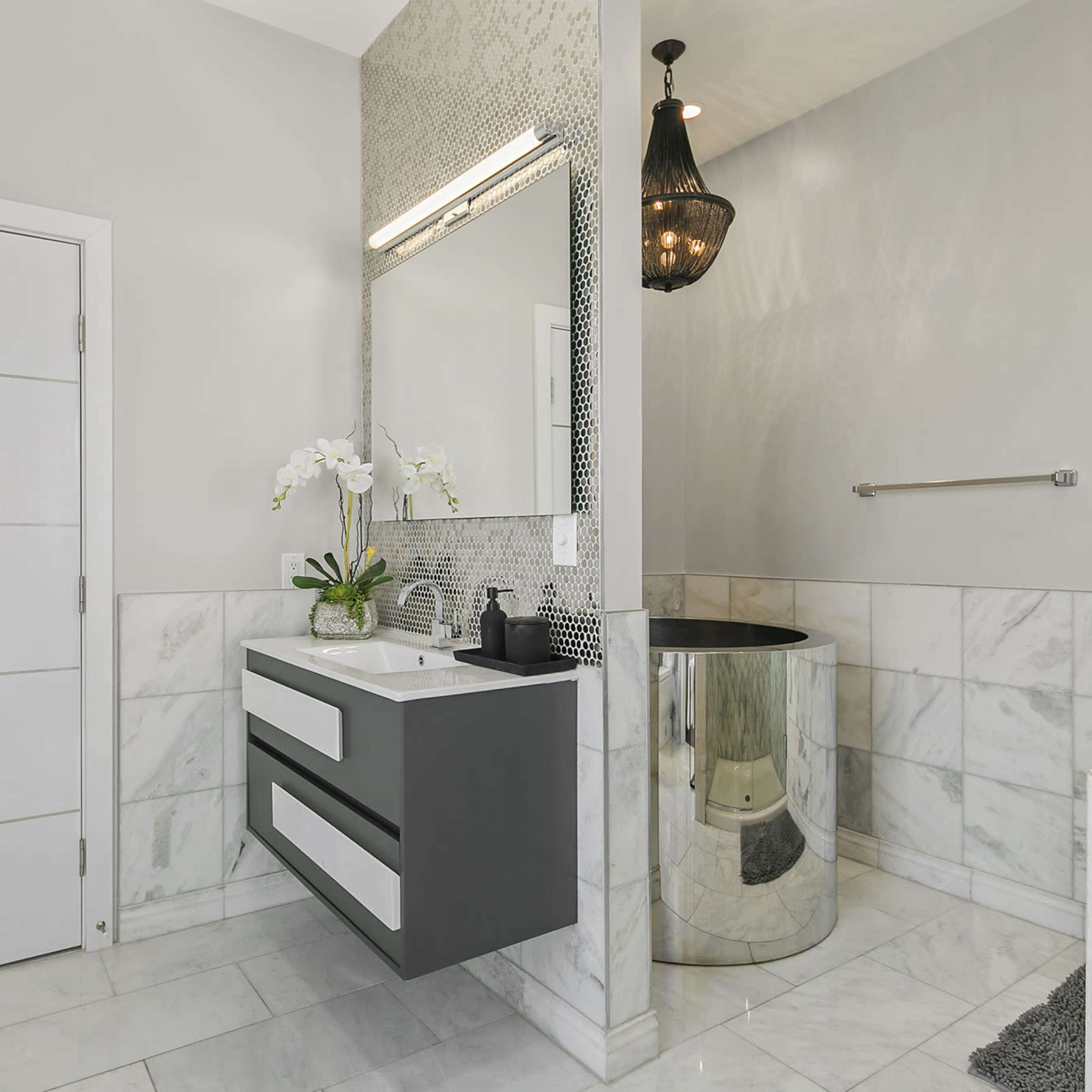 A modern bathroom features a mirror above a sleek gray vanity with two drawers, a round metallic stool, and a large walk-in shower area.