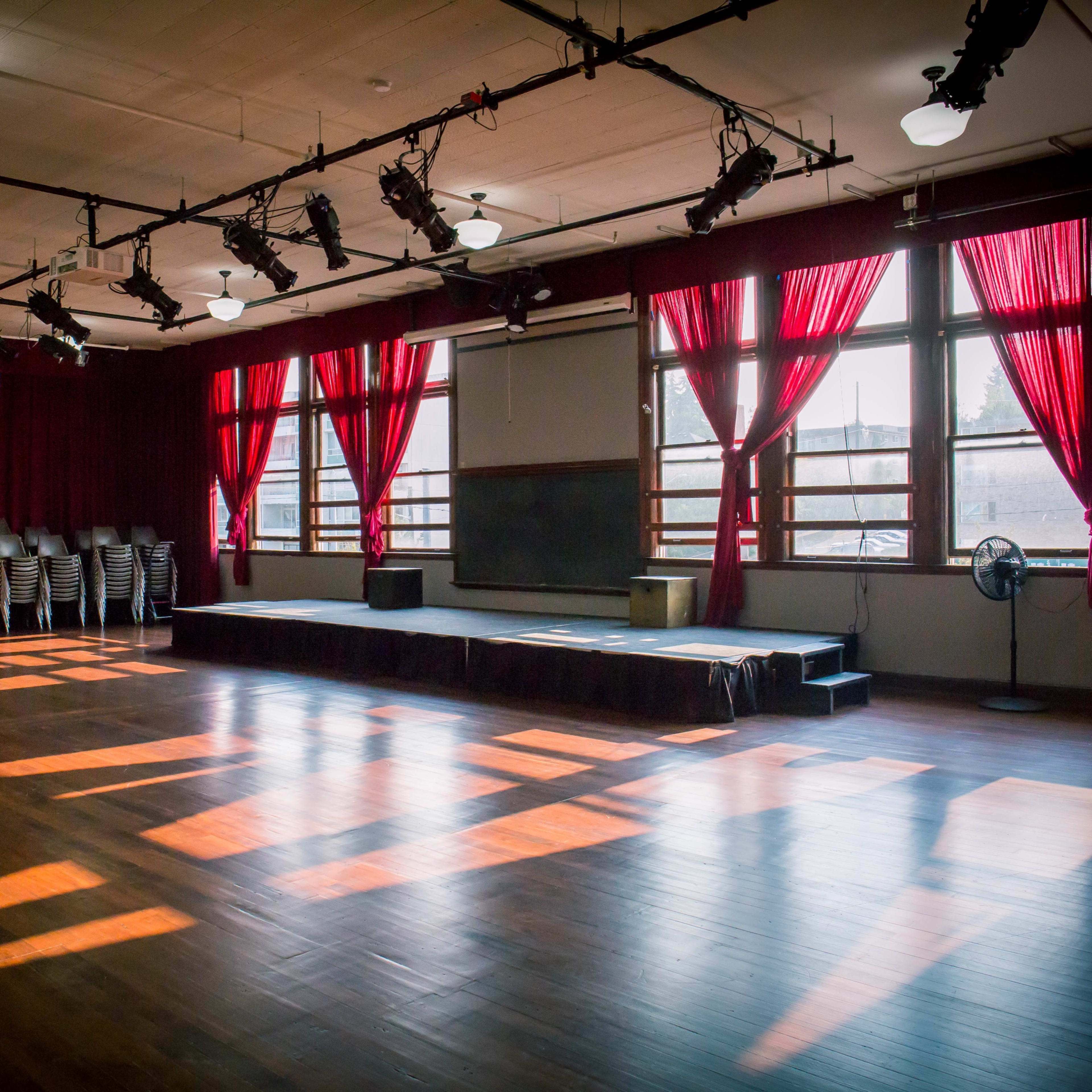 A spacious room with wooden floors, large windows draped in red curtains, and a small stage at the front.