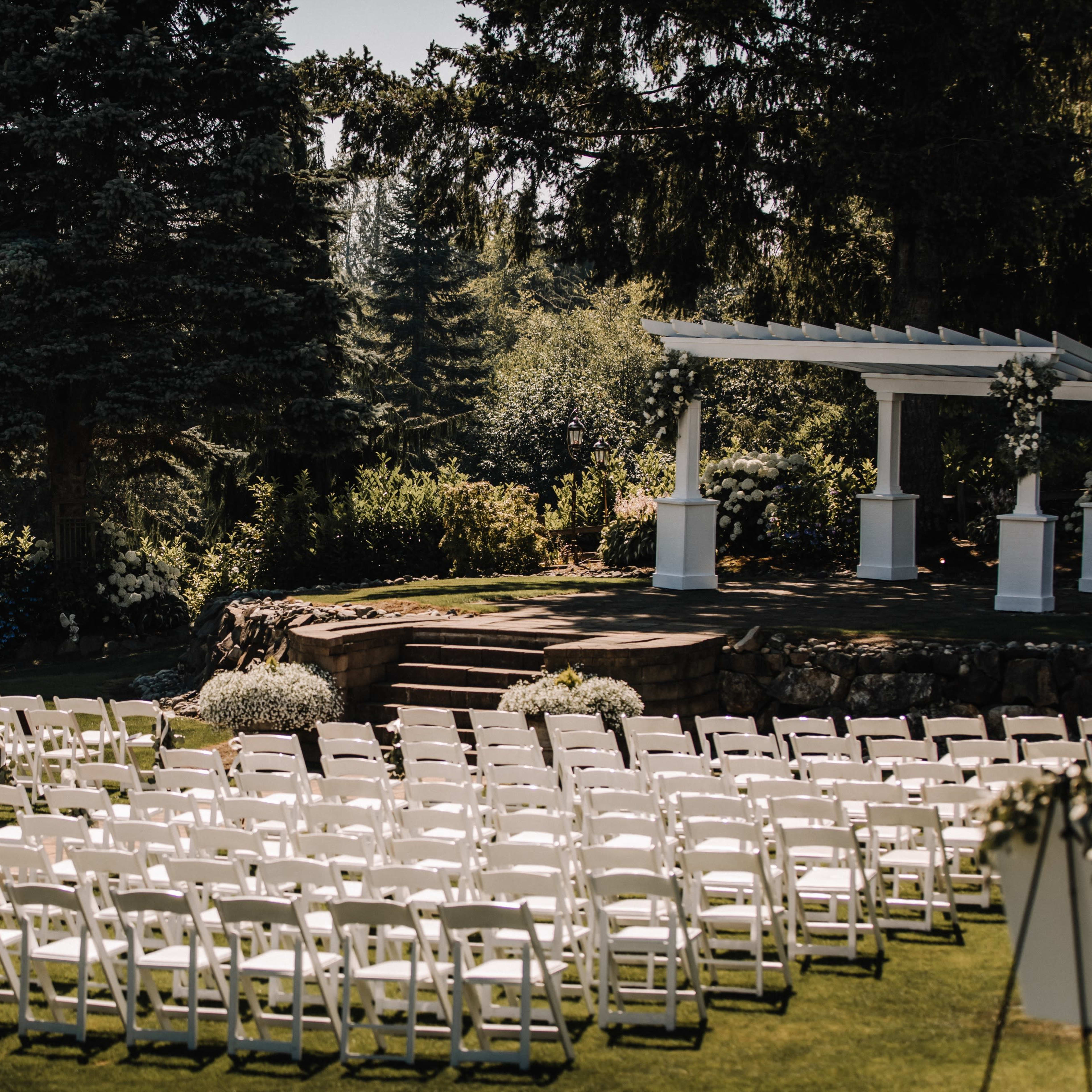 A large outdoor wedding venue features rows of white chairs arranged in front of a decorated arch on a sunny day.