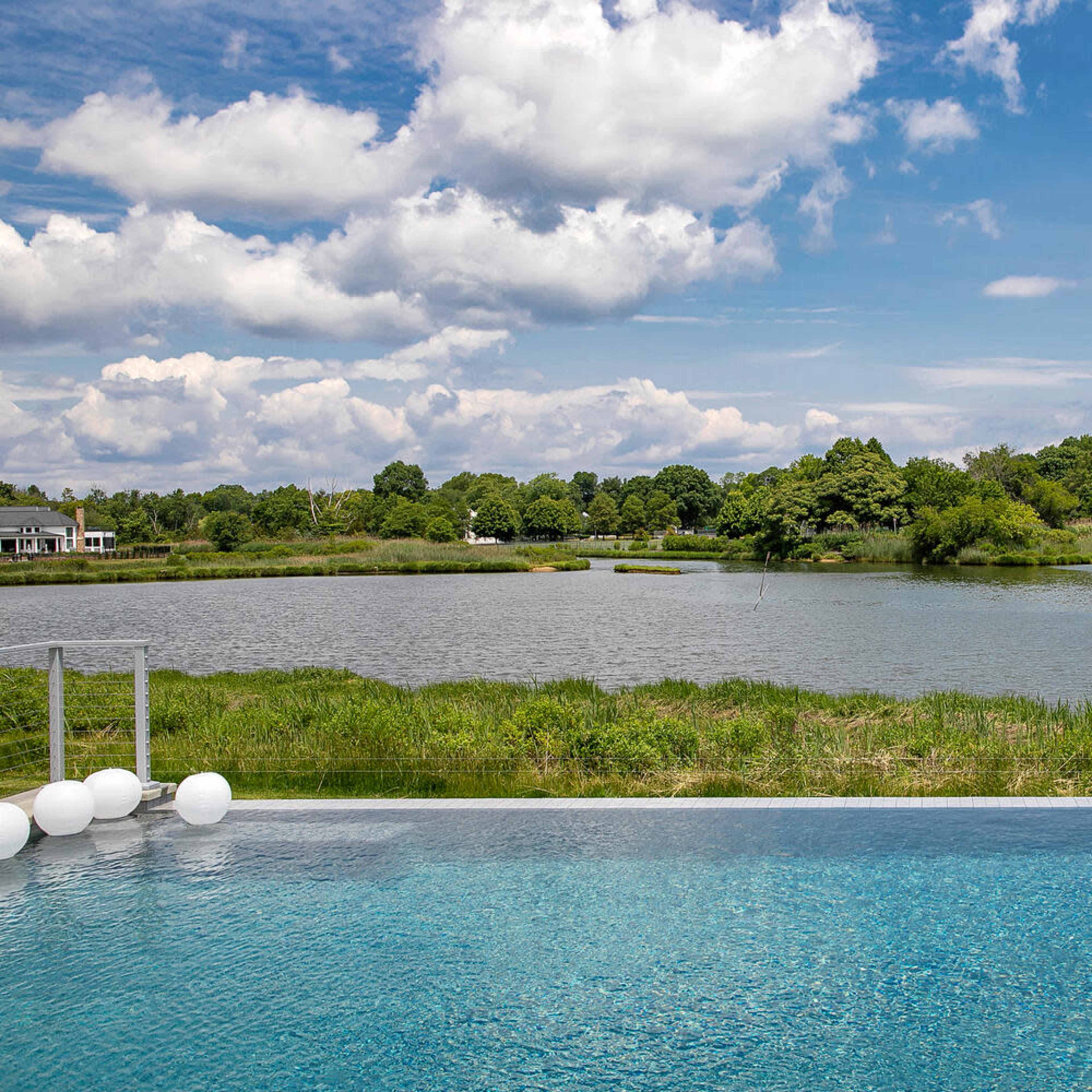 A clear view of a swimming pool overlooking a serene lake surrounded by lush greenery and a cloudy sky.