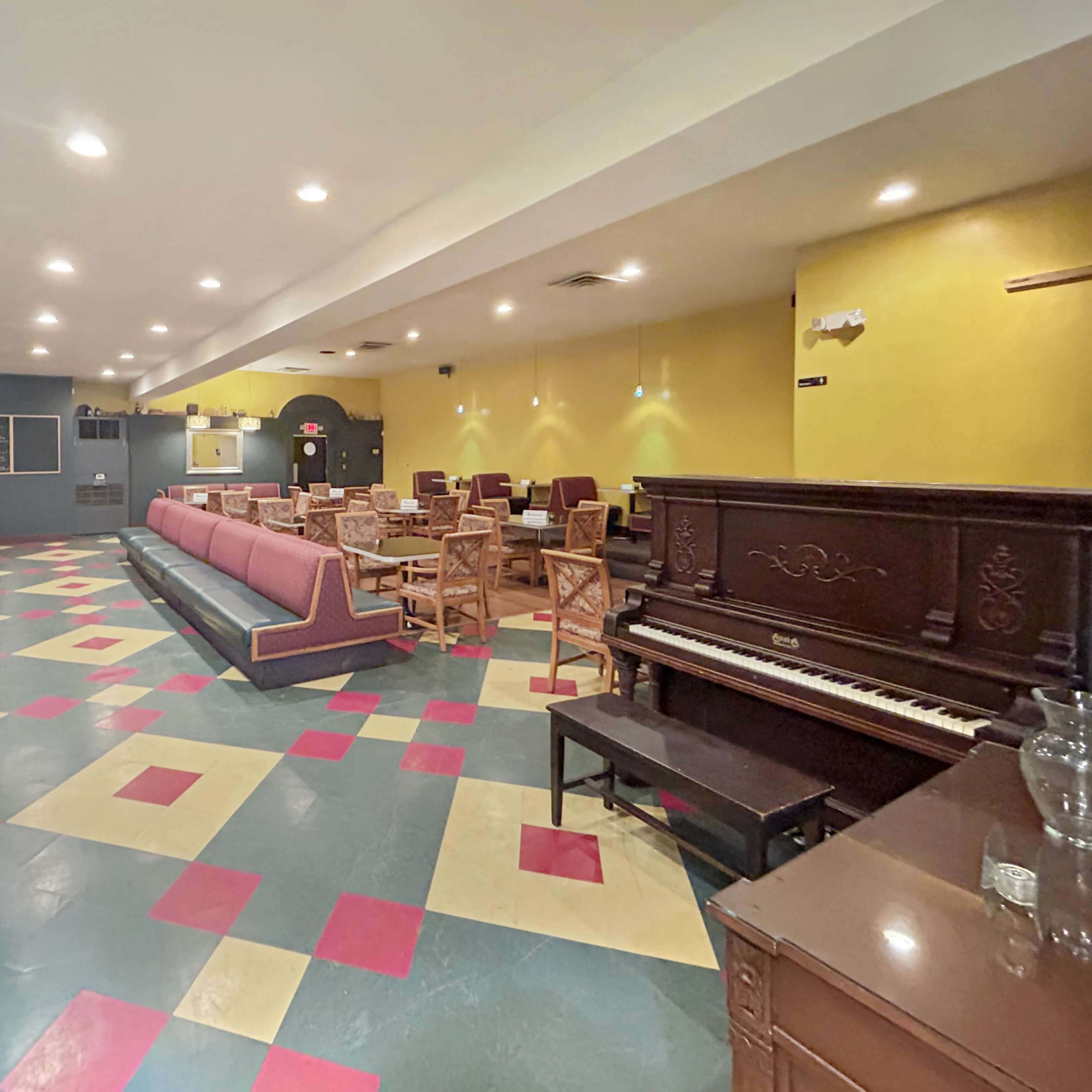 An empty café or lounge with colorful tiled flooring, a piano on one side, and arranged seating areas featuring wooden tables and chairs.