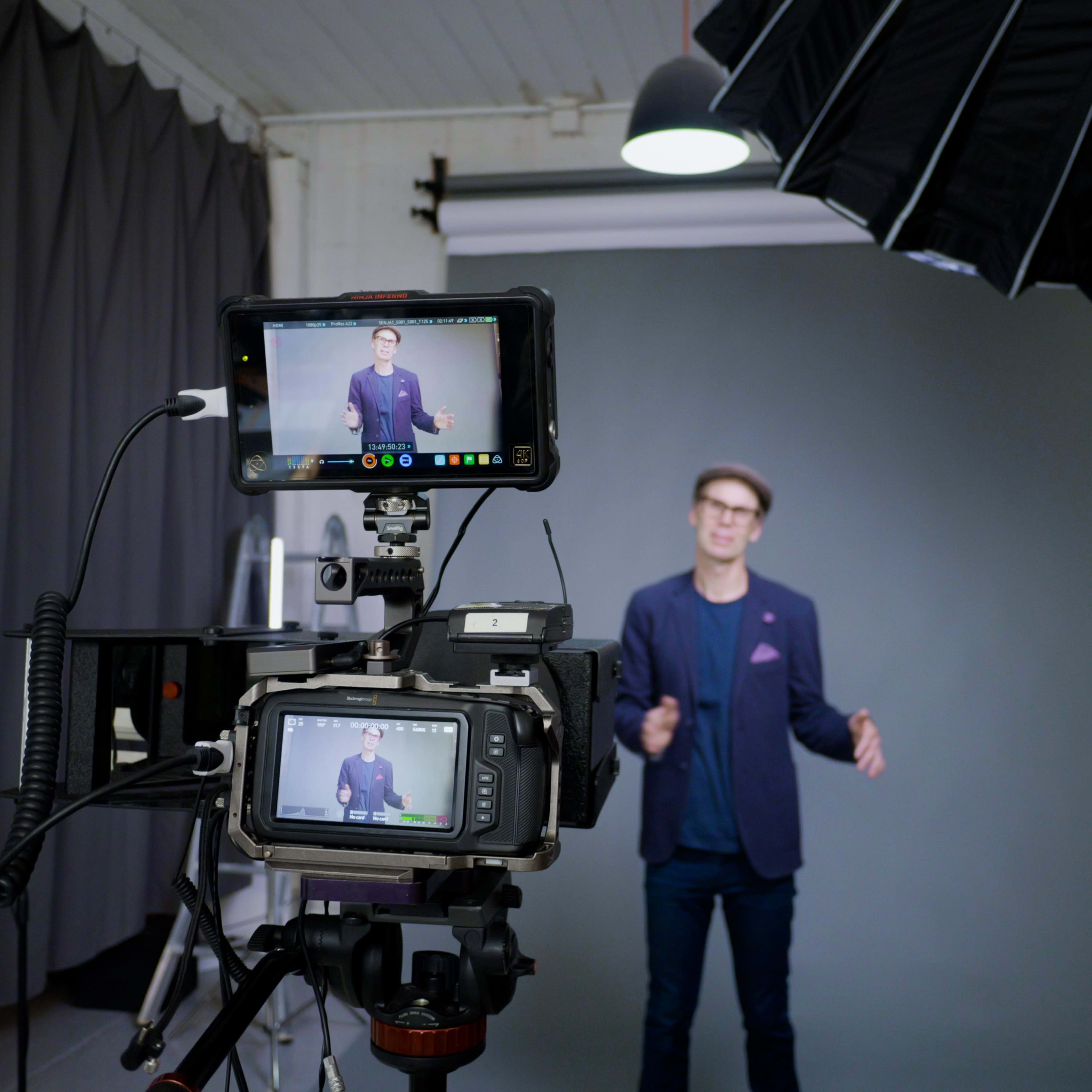 A man is standing in front of a gray backdrop while being filmed by a camera set up with monitors and lighting equipment.