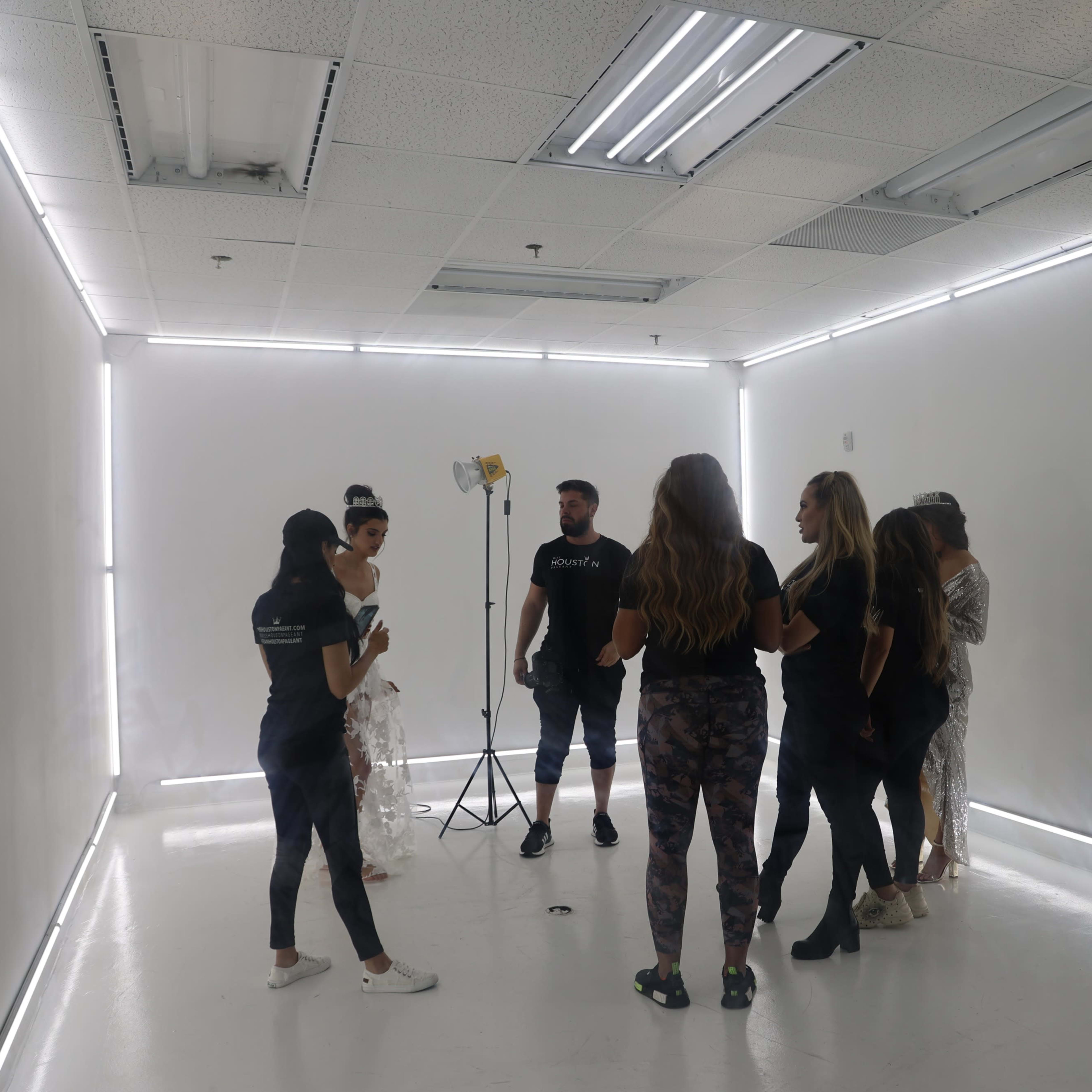 A group of seven people stands together in a brightly lit, minimalistic studio space with white walls and ceiling lights.