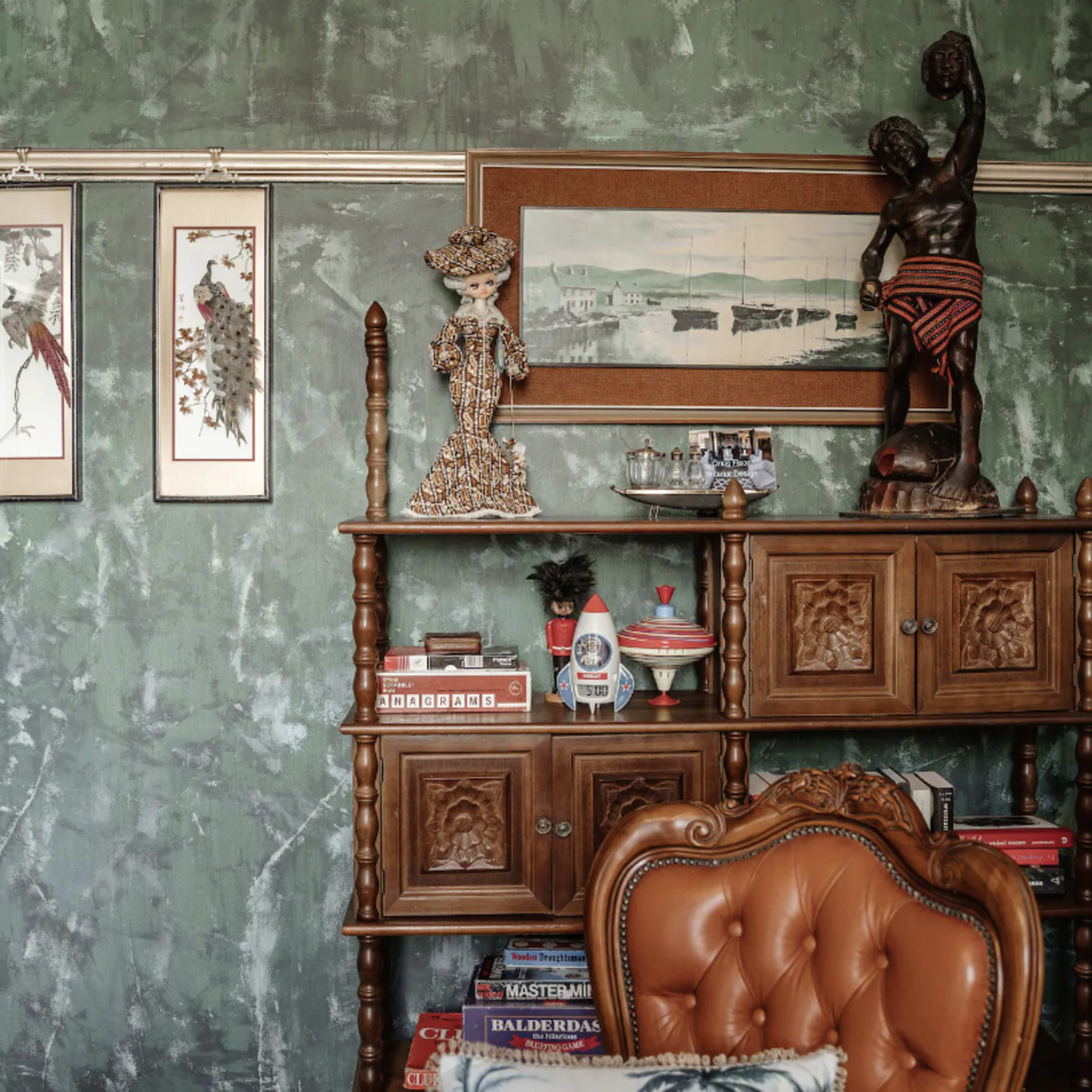 A wooden shelf adorned with decorative items, framed artwork, and a vintage chair against a textured green wall.