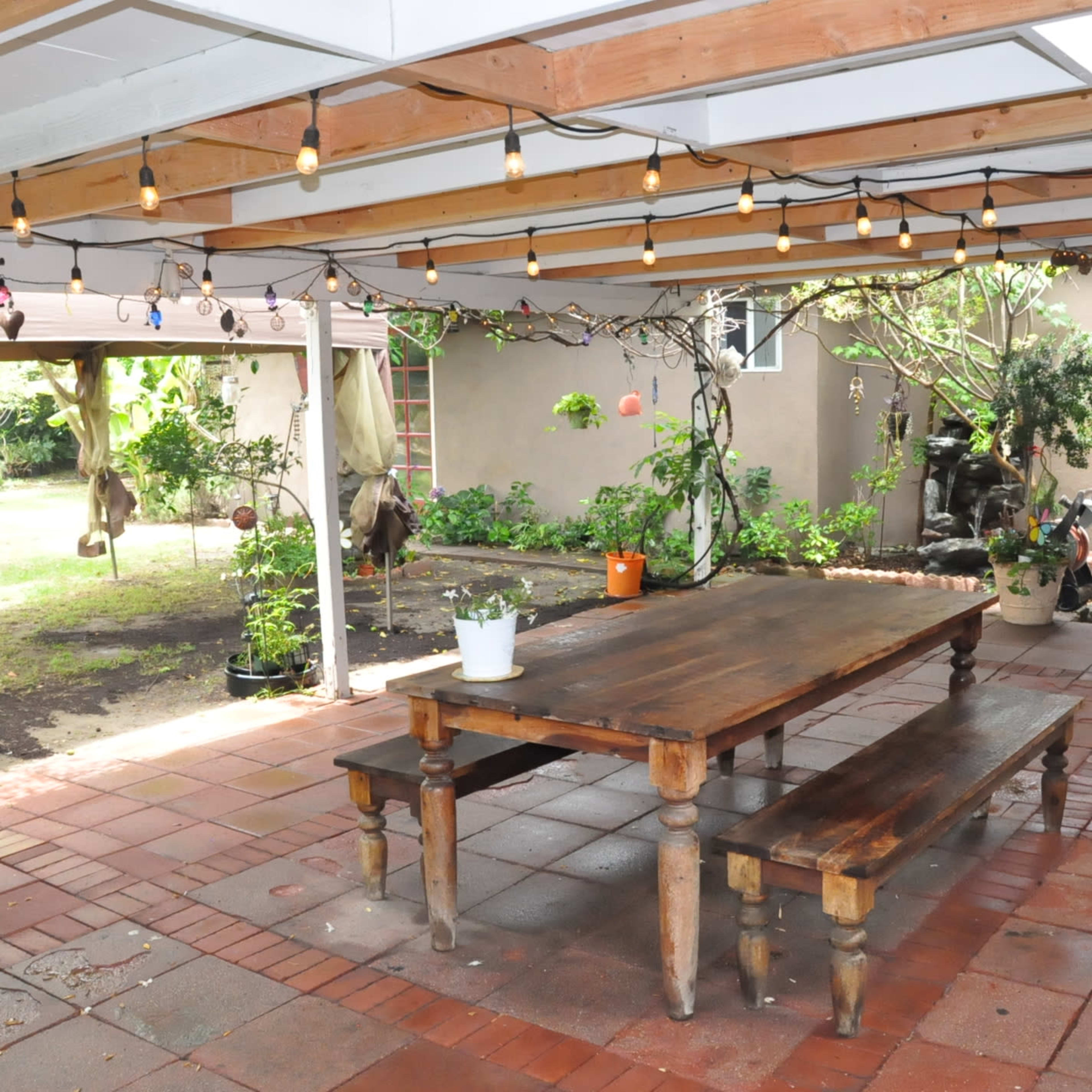 A covered outdoor patio area featuring a wooden table and benches, with string lights overhead and green plants in the background.