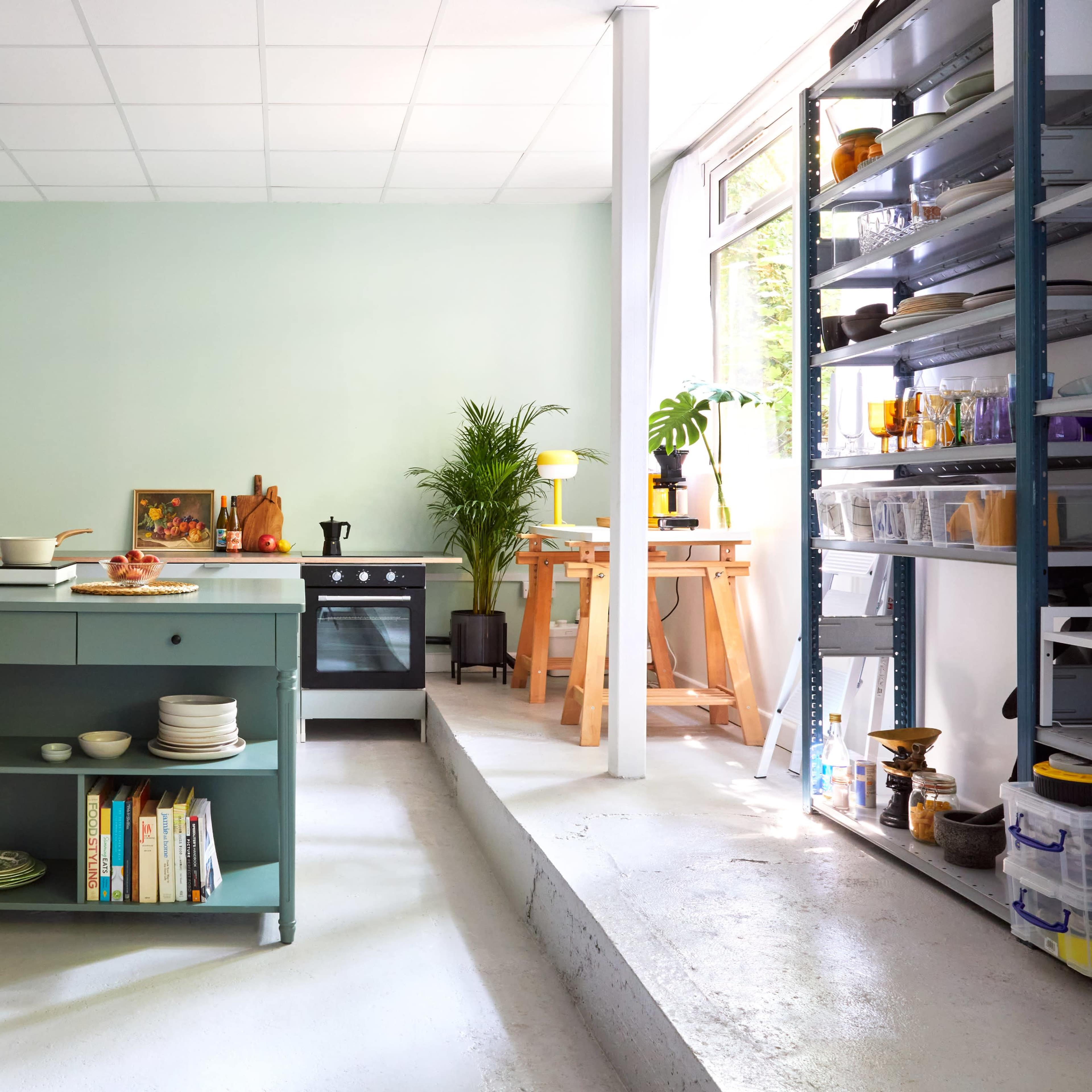 A modern kitchen features a central island with a turquoise base, white countertops, and shelves stocked with dishes, surrounded by large windows and various plants.