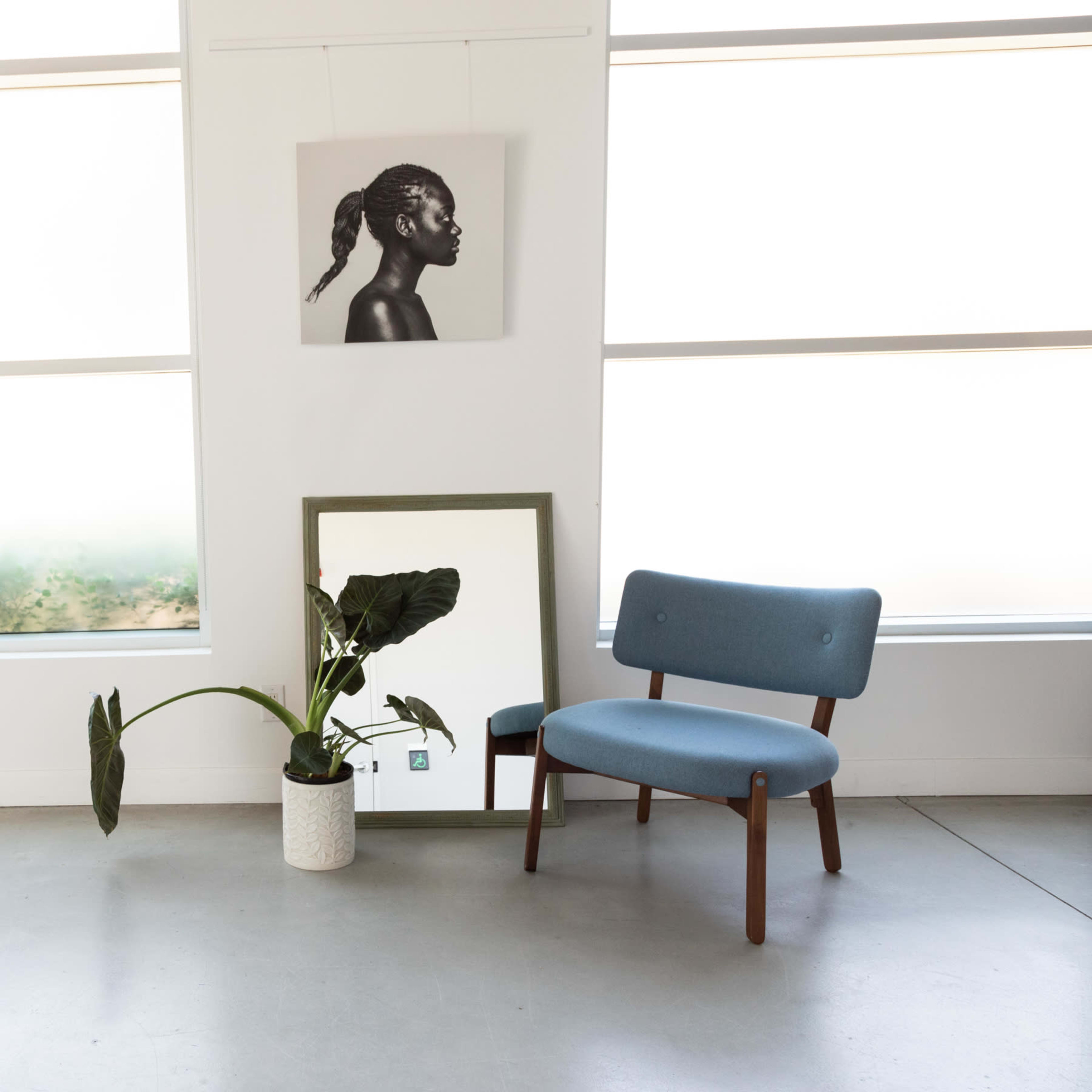 A blue chair is positioned beside a potted plant and a mirror in a brightly lit room with large windows.