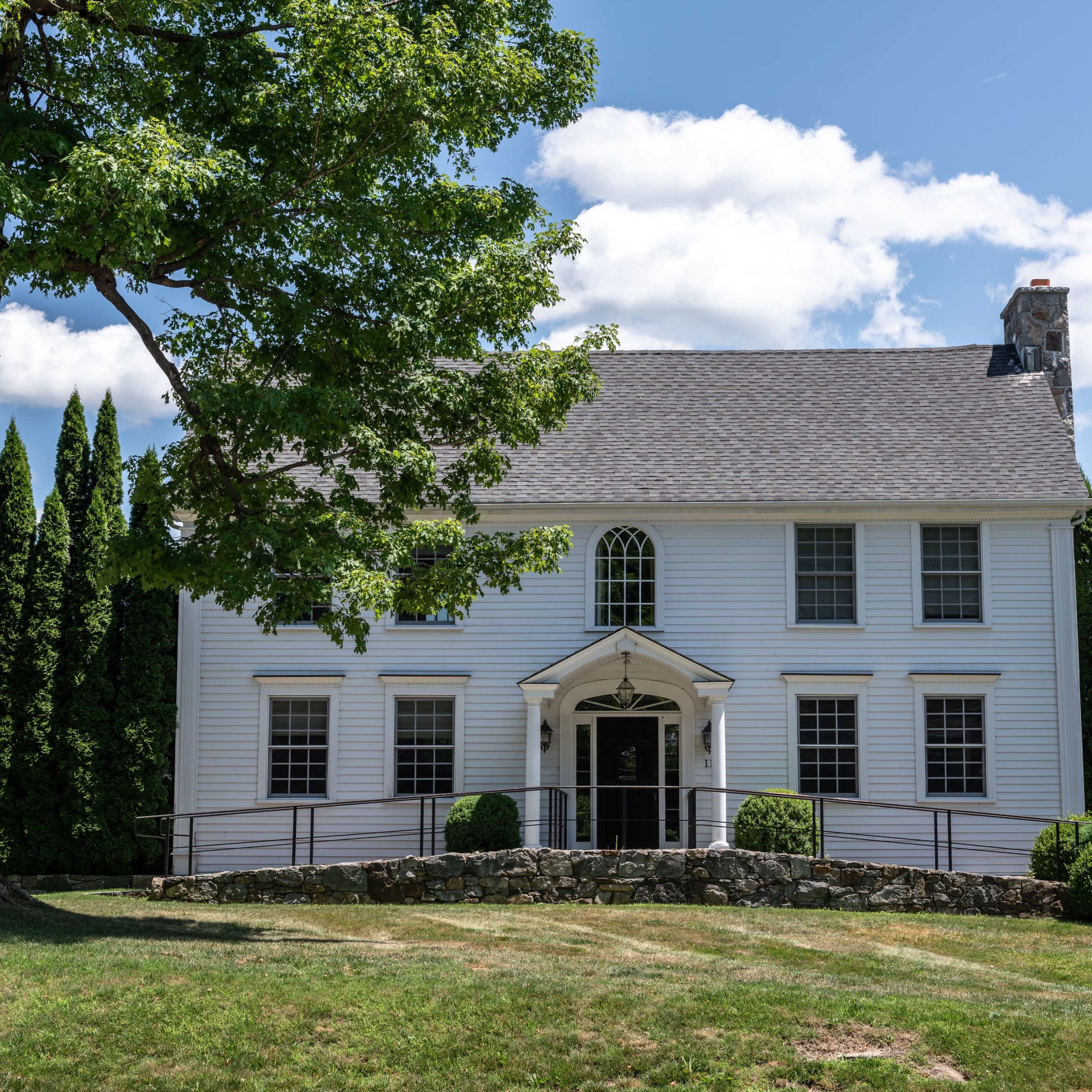 A two-story white house with a gray roof and a front porch is surrounded by well-maintained grass and tall evergreen trees.