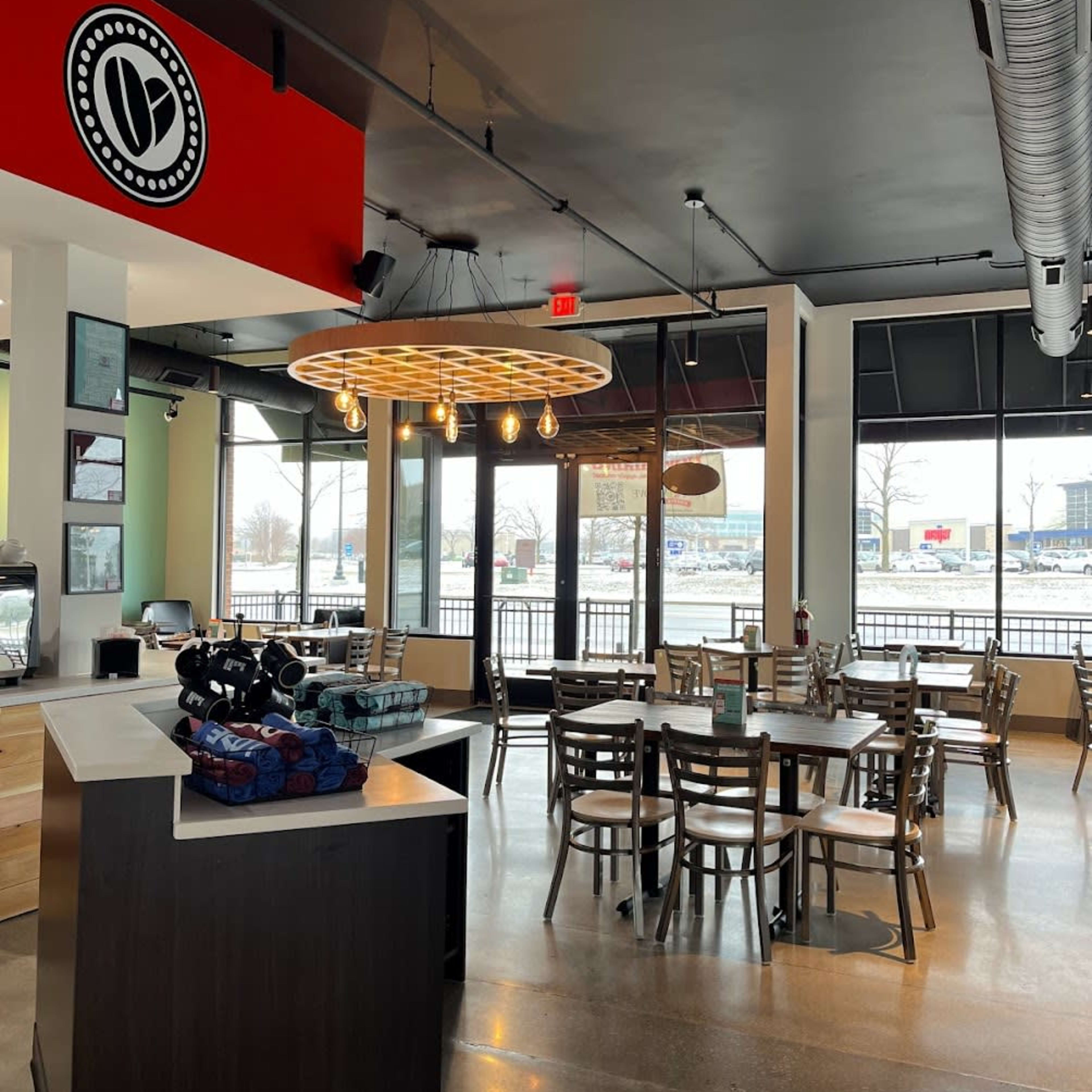 A modern café interior with a round light fixture, wooden tables and chairs, and large windows overlooking a view outside.