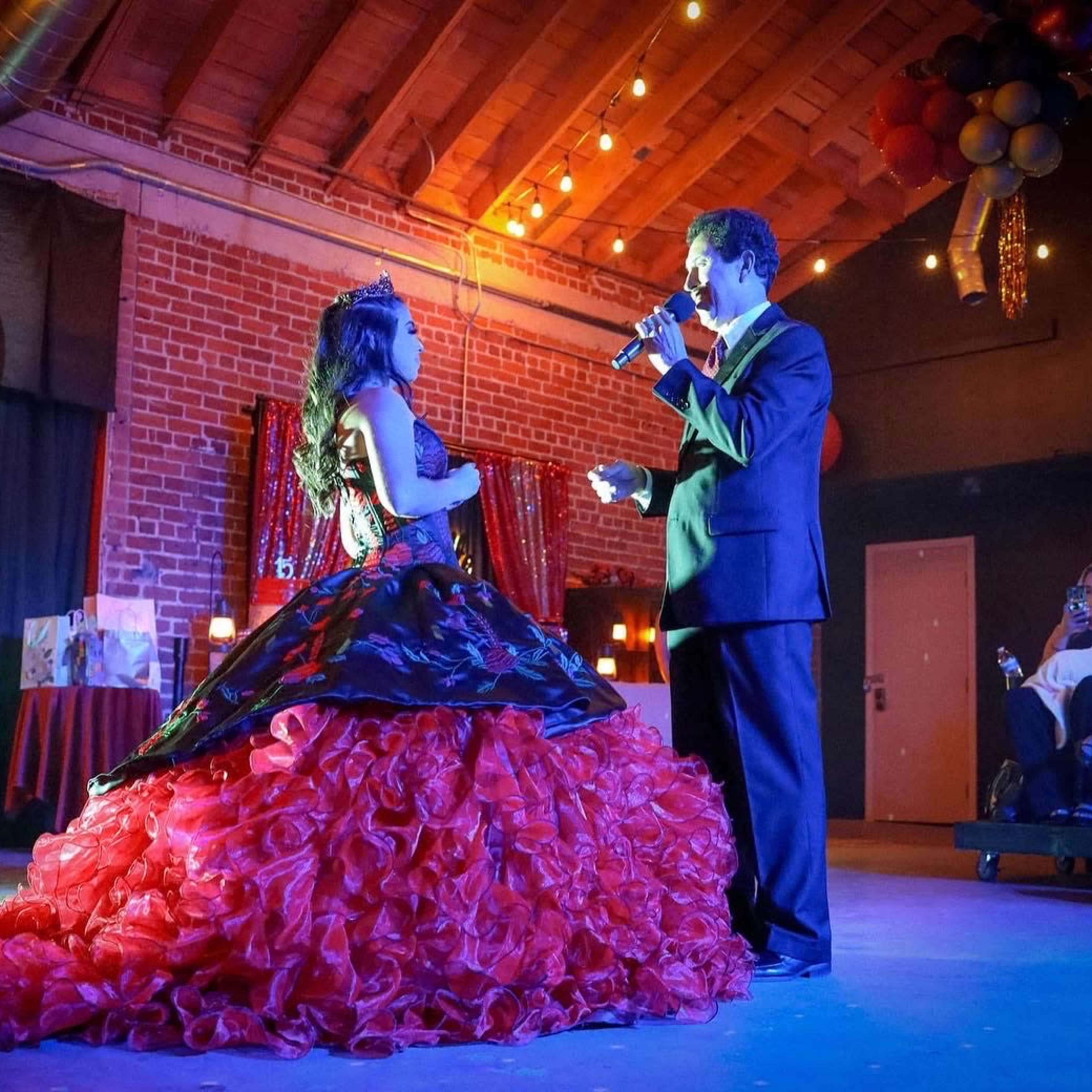 A young woman in a red and black gown stands on a stage while a man in a suit sings to her in a decorated event space.