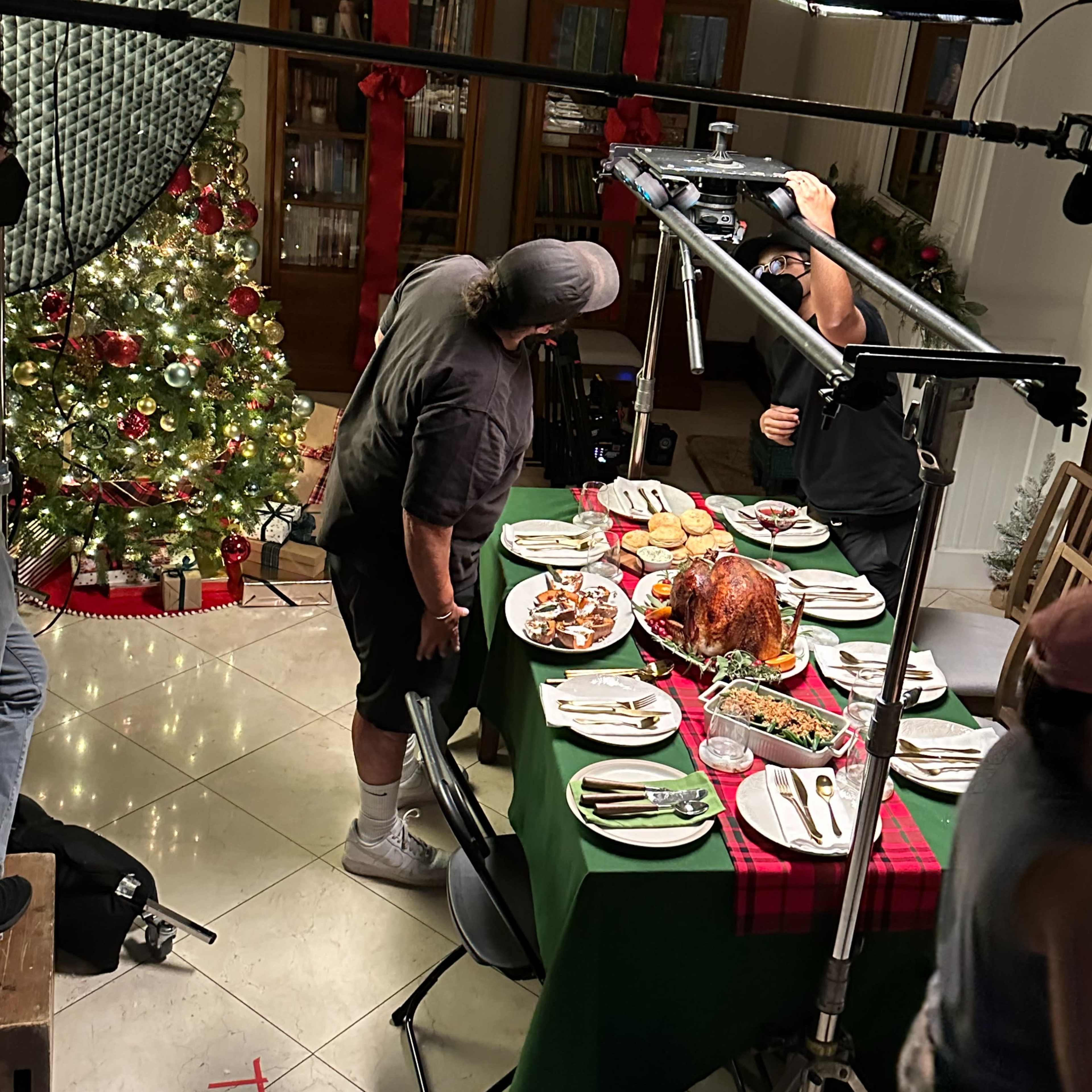 A crew sets up lighting for a dinner scene featuring a decorated table with a turkey and various dishes, surrounded by festive decorations and a Christmas tree.