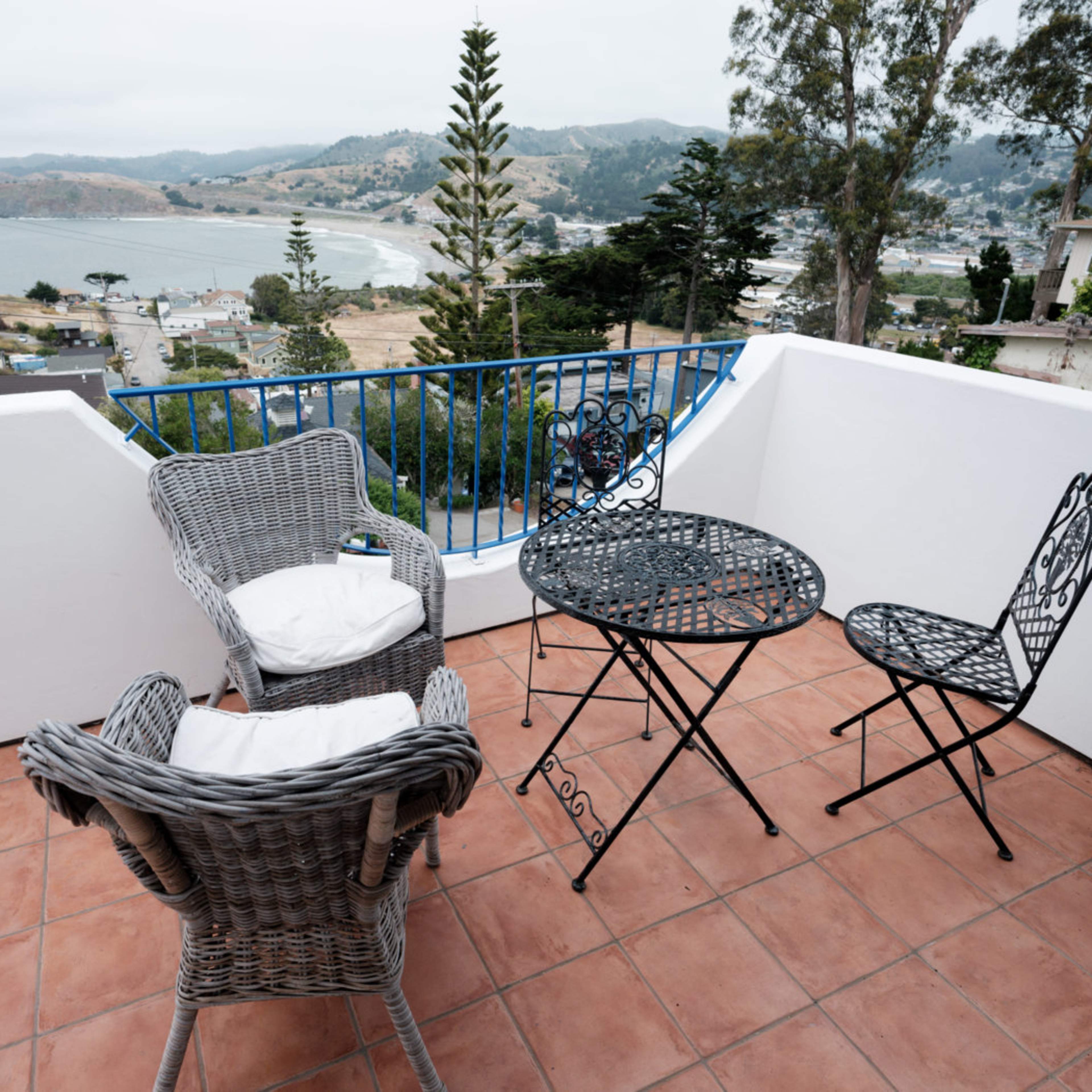 A small balcony features a round metal table and two chairs, with a view of a coastal landscape in the background.