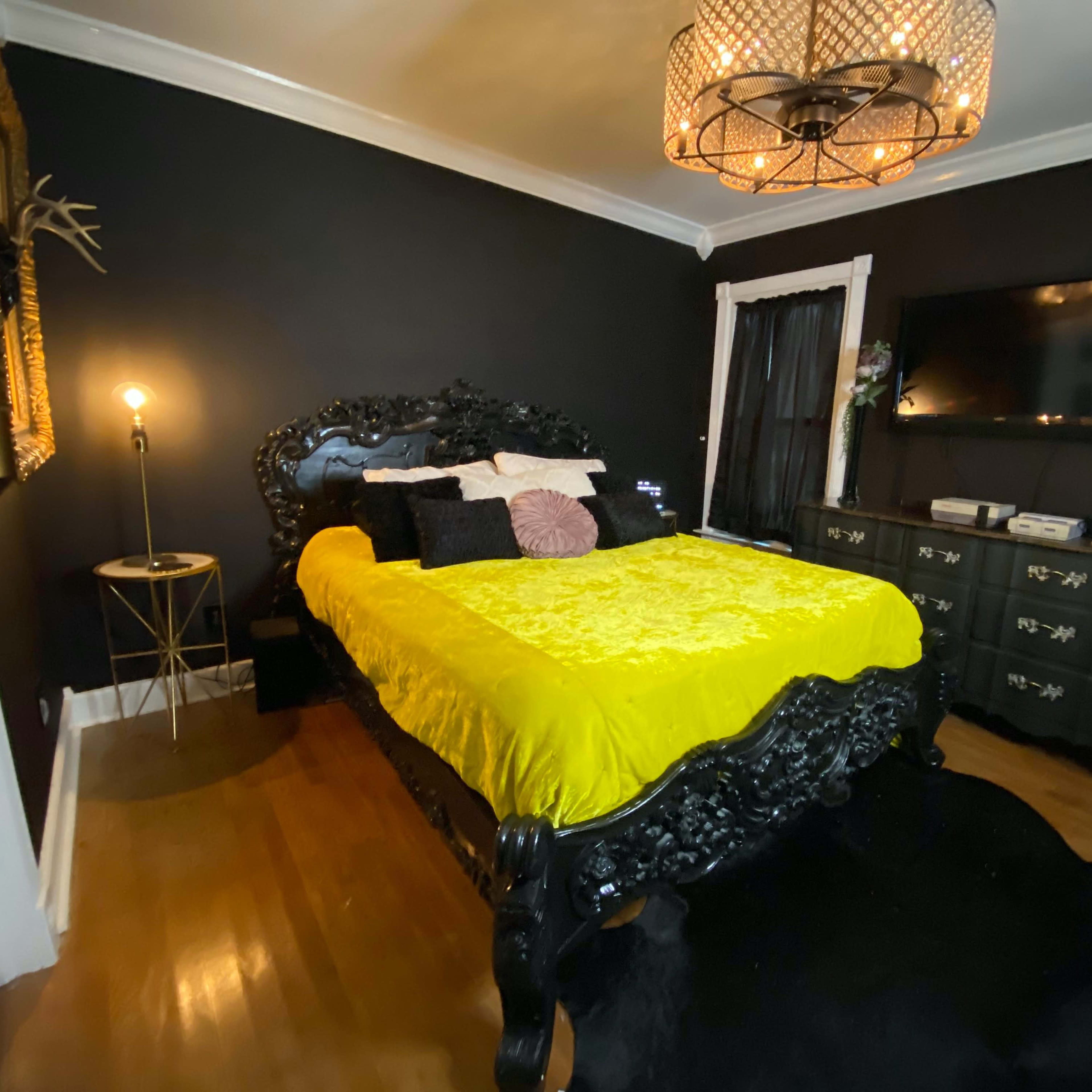 The bedroom features a black walls, a decorated yellow bedspread, a black ornate bed frame, and a chandelier overhead.