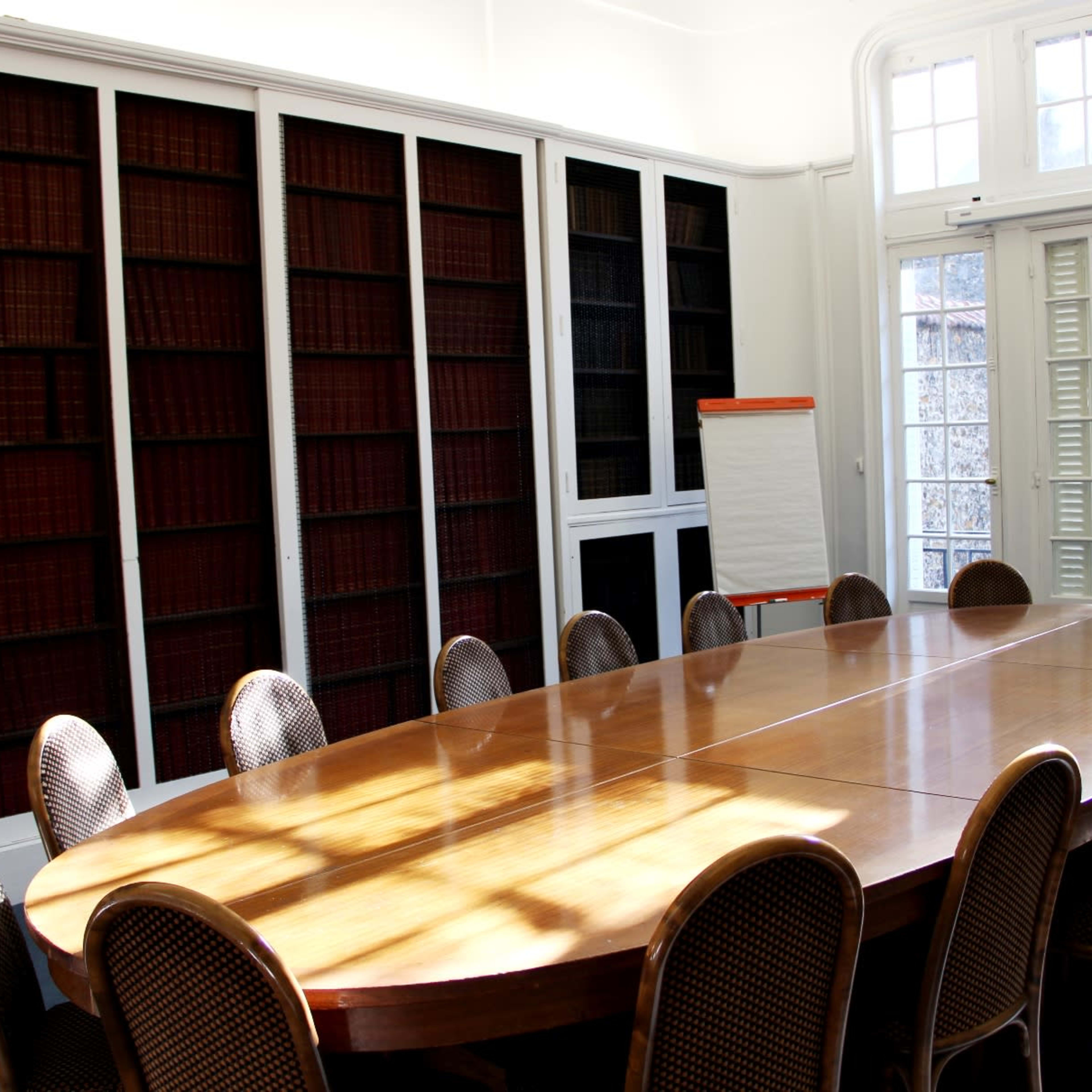 A wooden oval conference table surrounded by chairs in a bright room with bookshelves and large windows.
