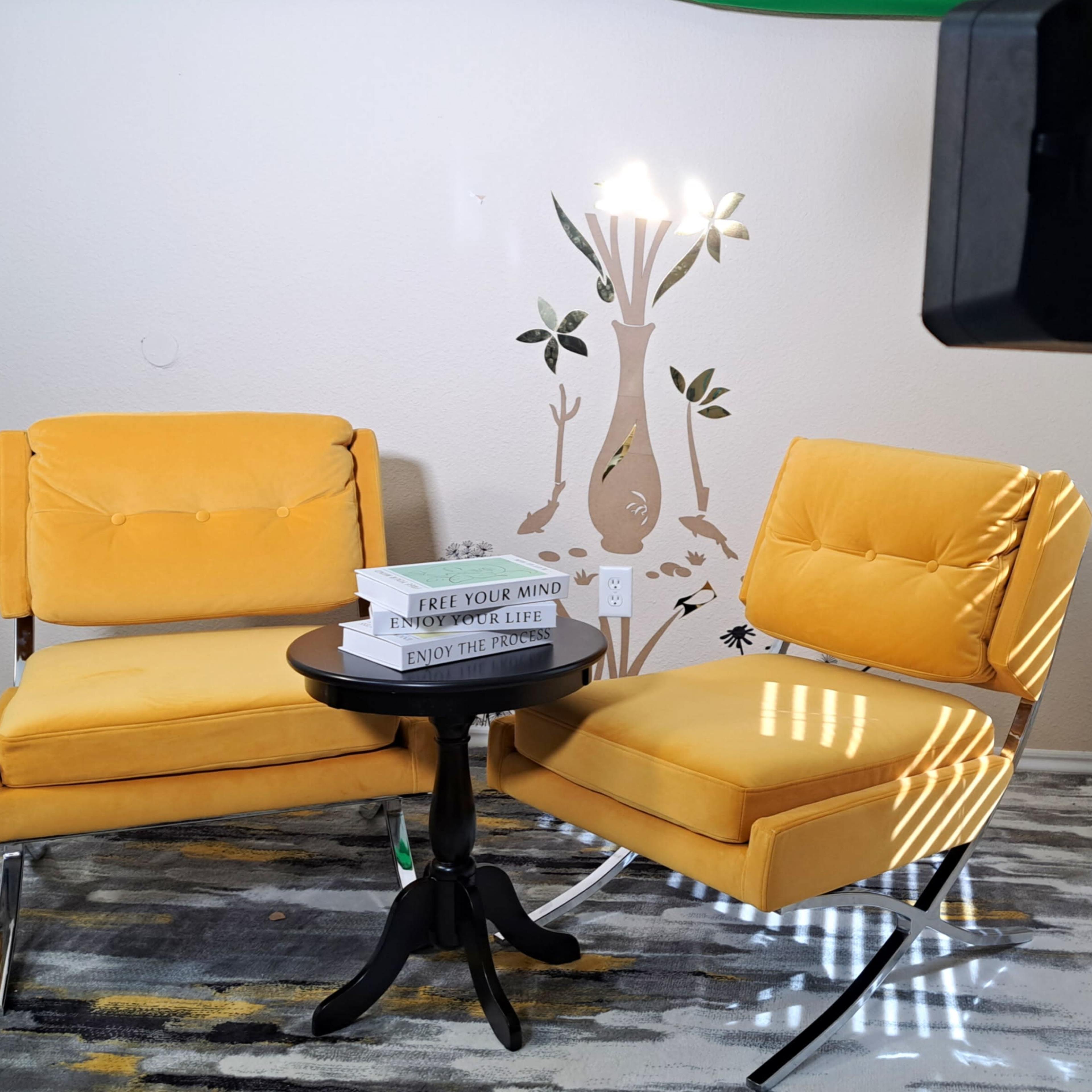 A cozy interior scene featuring two yellow chairs, a small black table with books, and a decorative wall mural of a tree.