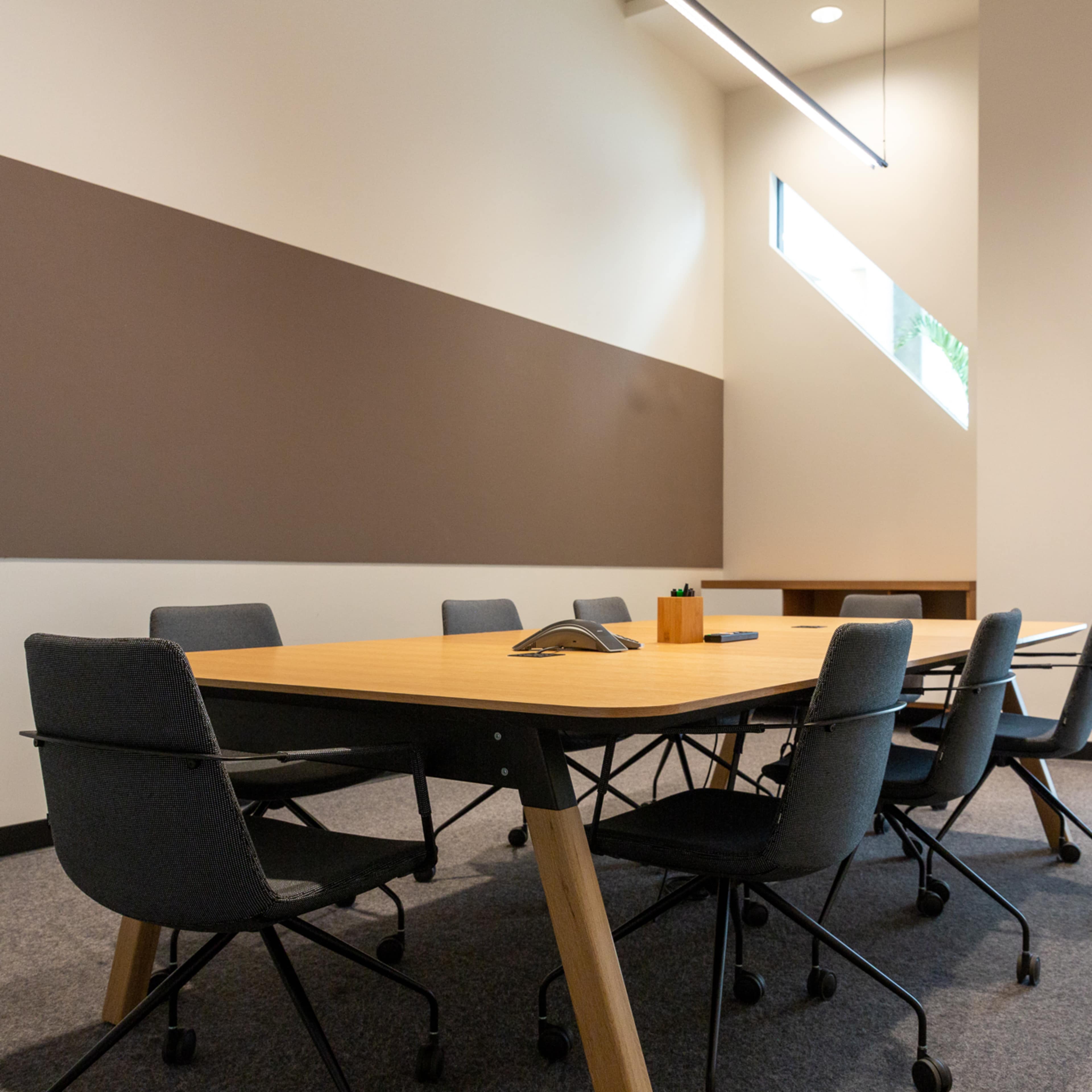 A conference room features a large wooden table surrounded by rolling chairs, with a doorway leading to an adjacent space.