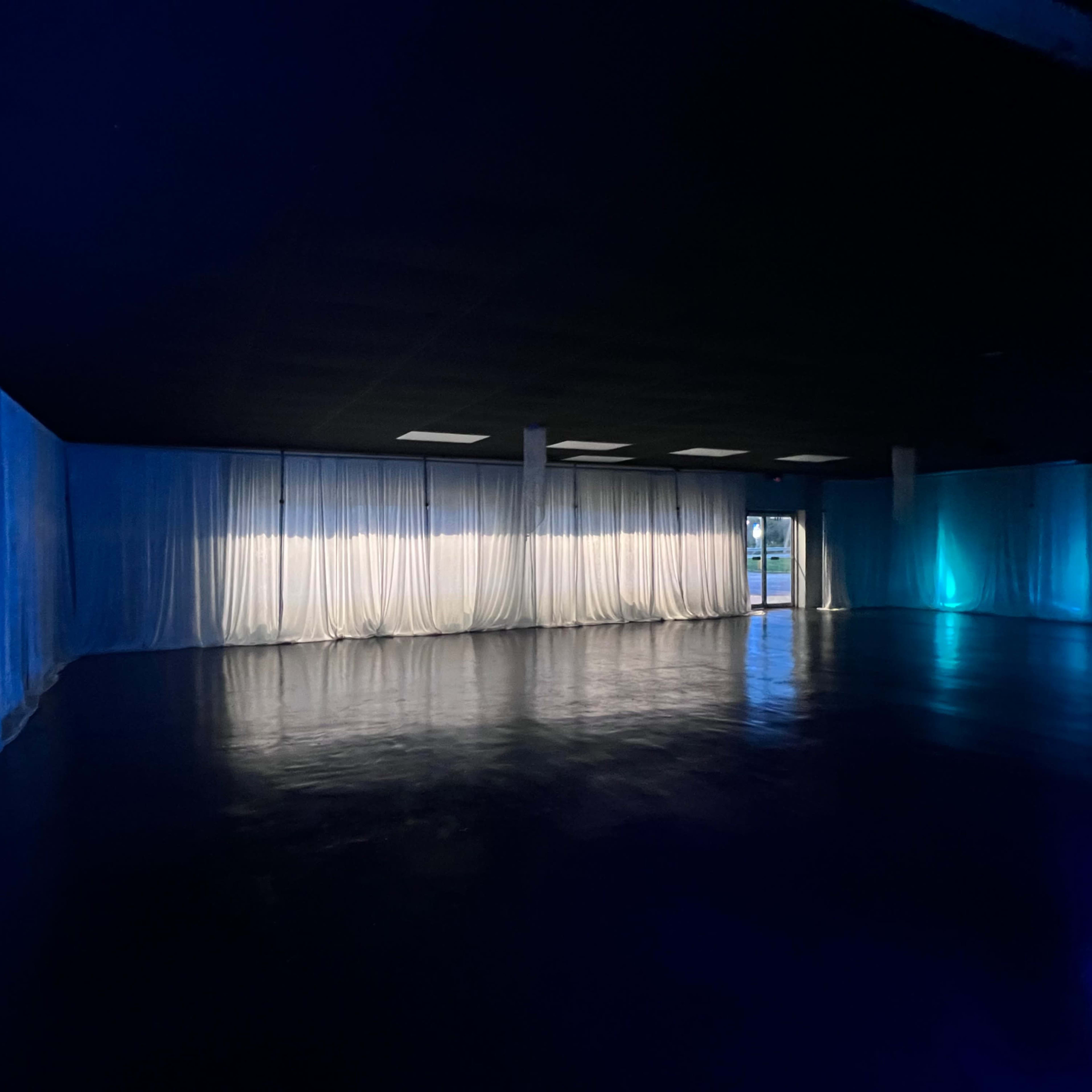 An empty room with dark flooring and blue-lit sheer curtains lining the walls.