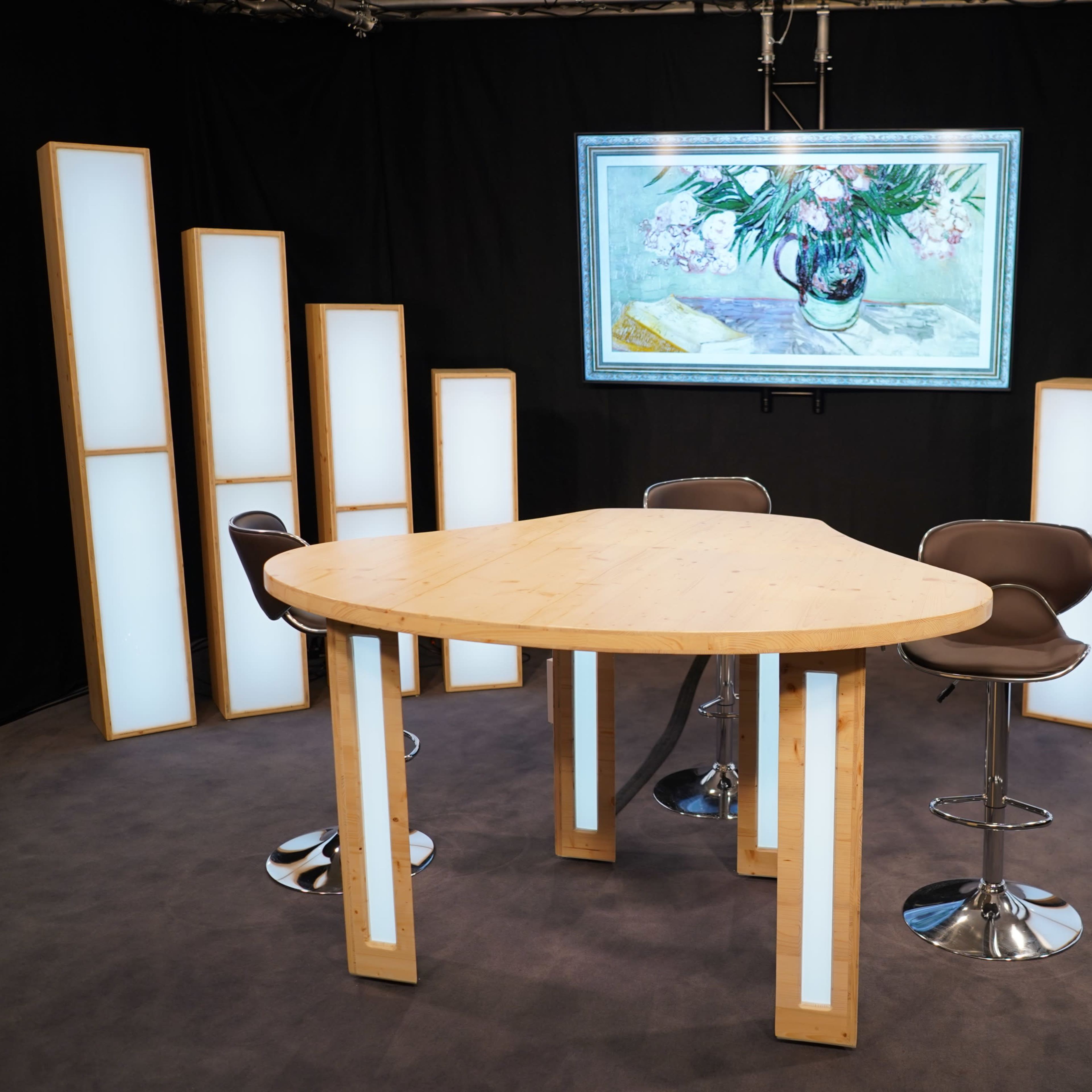A modern studio setup features a triangular wooden table surrounded by high stools, illuminated by tall, backlit panels and a large artwork displayed above.