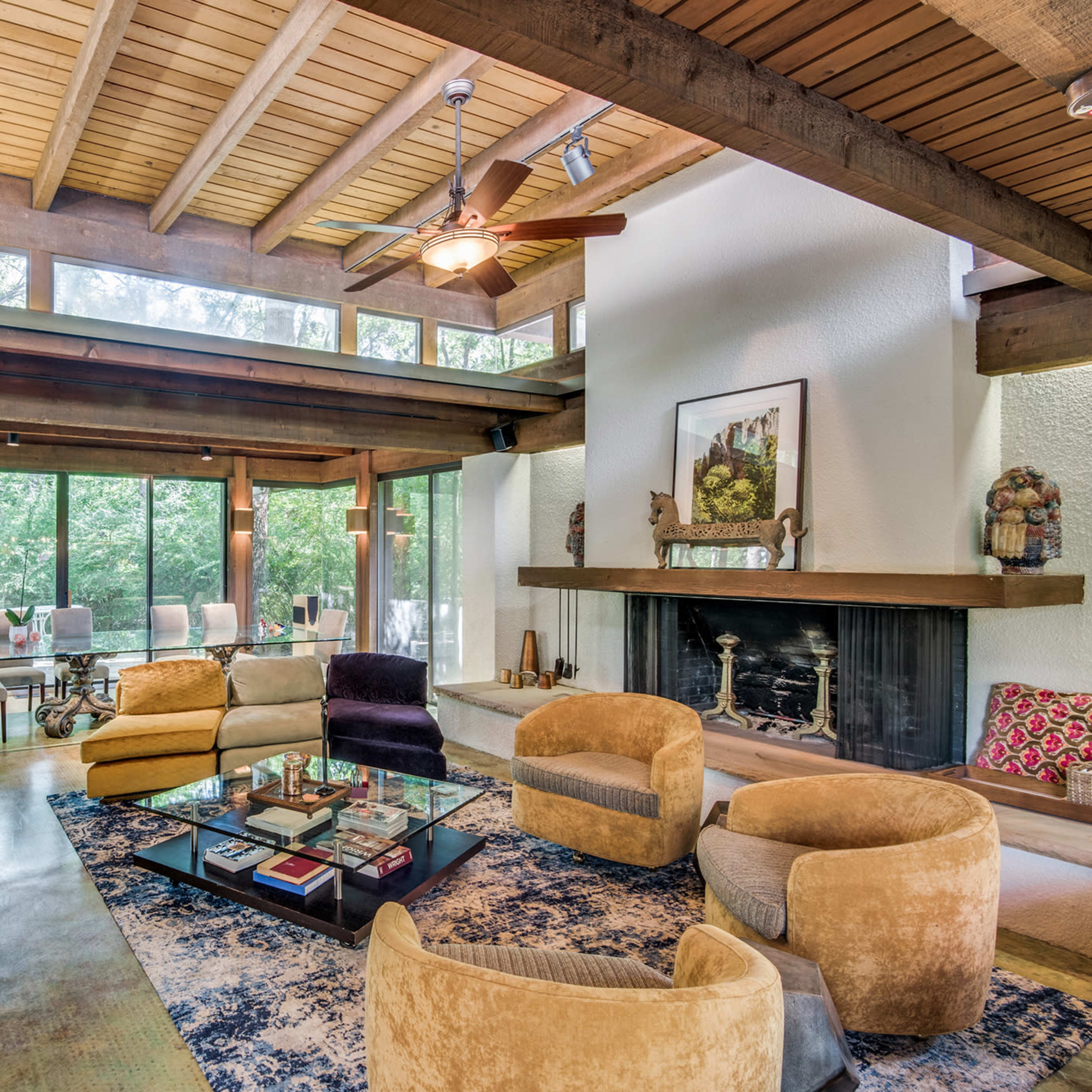 A modern living room with wooden beams, a stone fireplace, and large windows that overlook a wooded area.