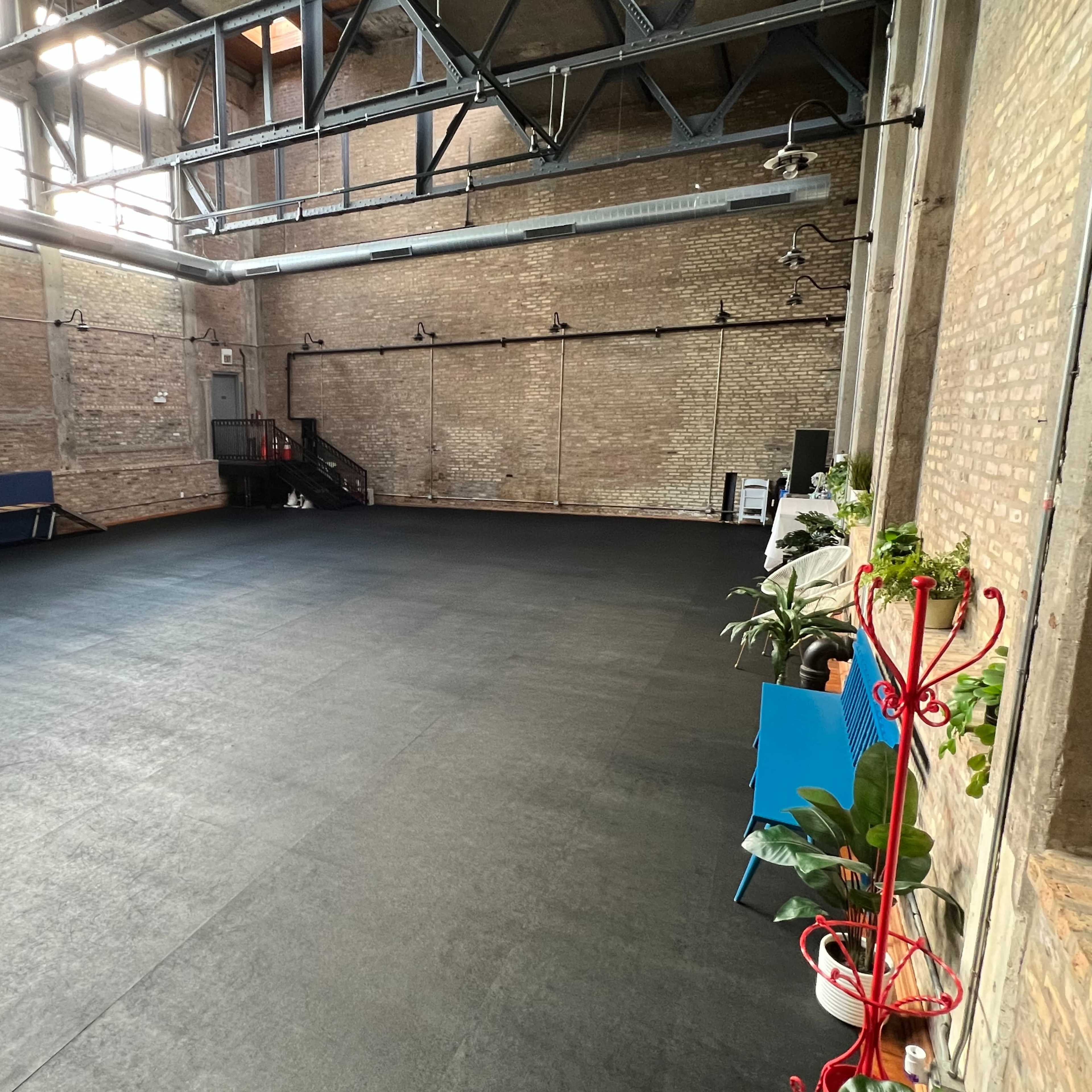 A spacious, empty indoor area with black rubber flooring and exposed brick walls, featuring a few pieces of furniture and plants along the edges.