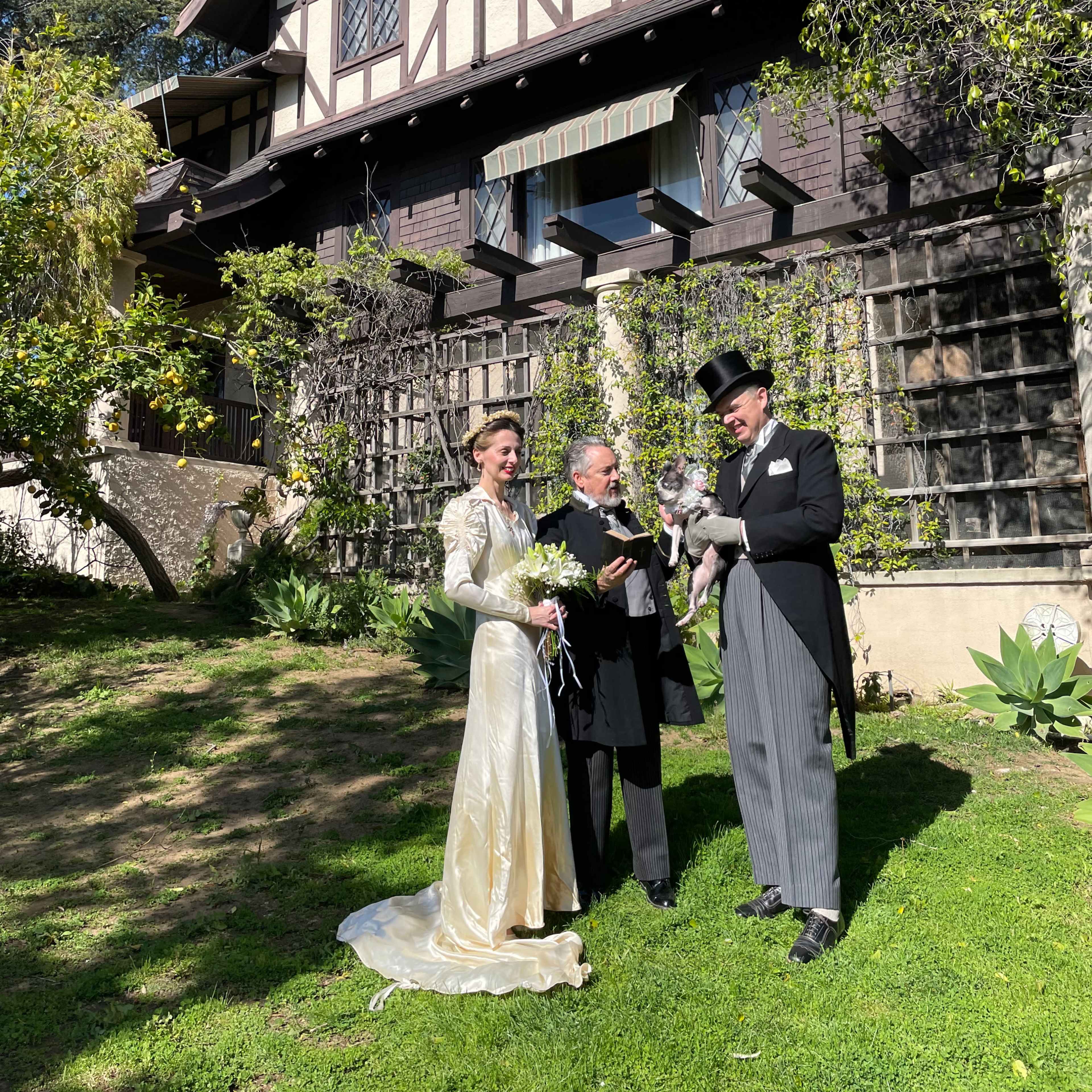 A bride in a white gown and a groom in formal attire stand with a man in a black coat in front of a large, multi-story house surrounded by greenery.