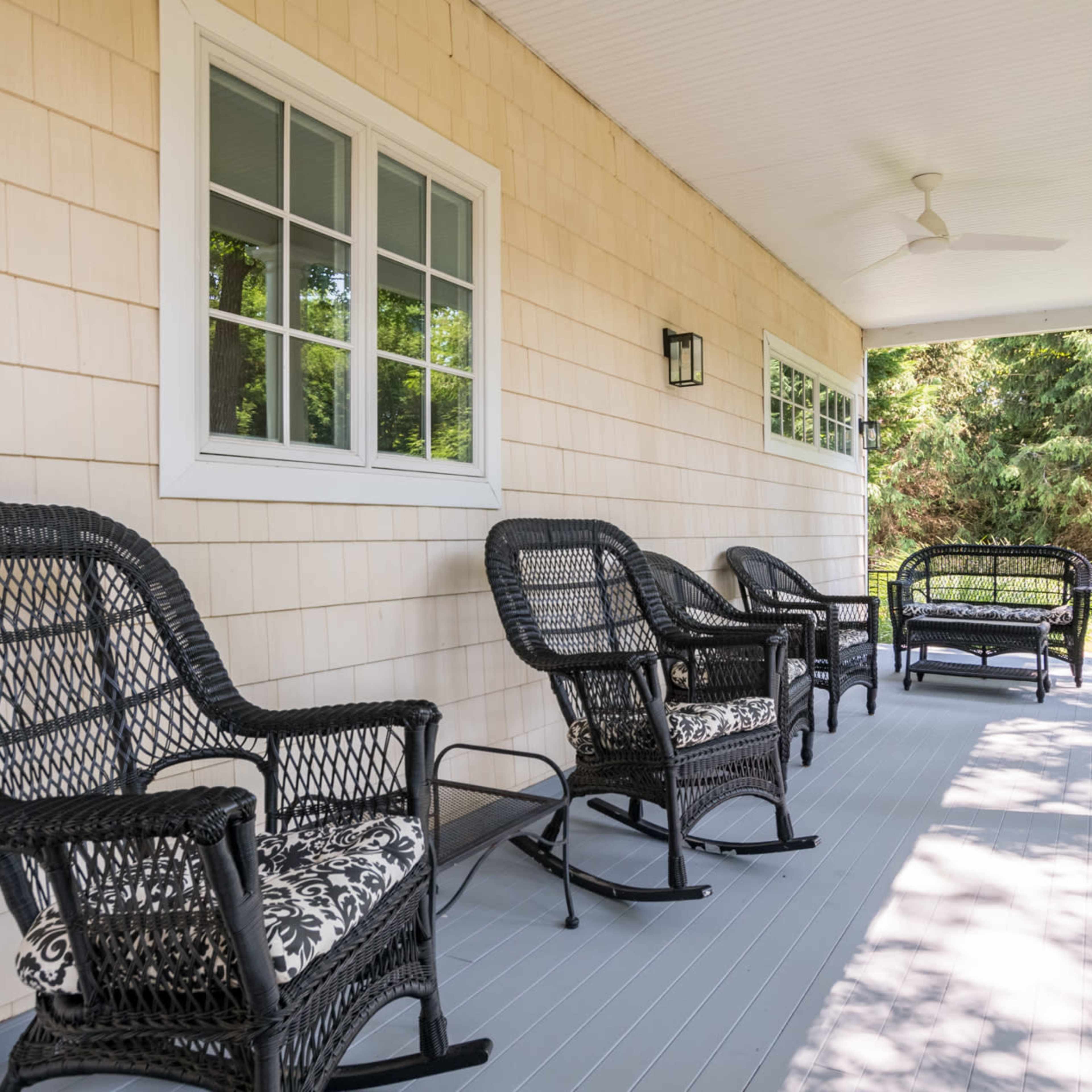 A porch with four black wicker chairs and a ceiling fan, surrounded by greenery.