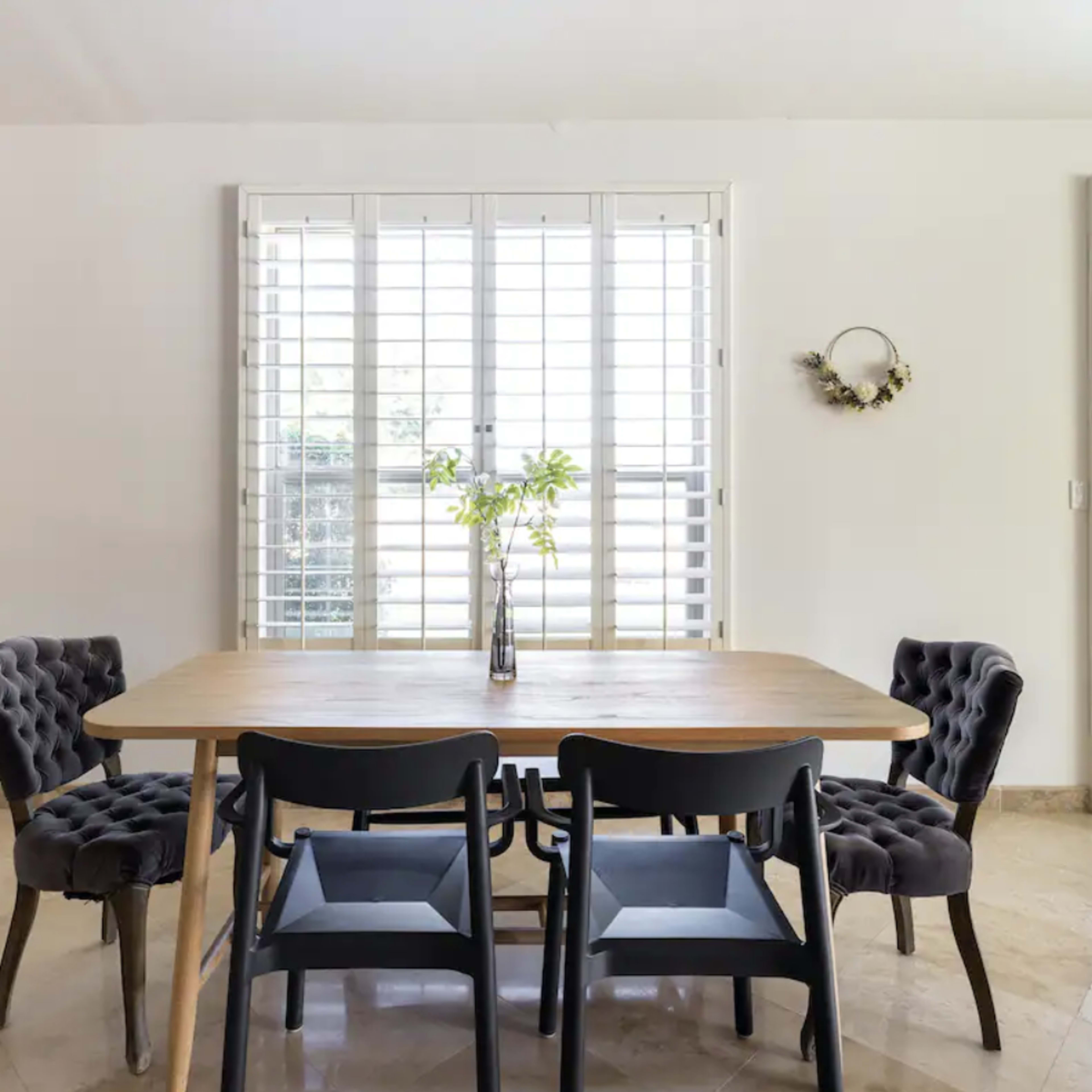 A modern dining area features a wooden table surrounded by upholstered chairs, with light filtering through shuttered windows.