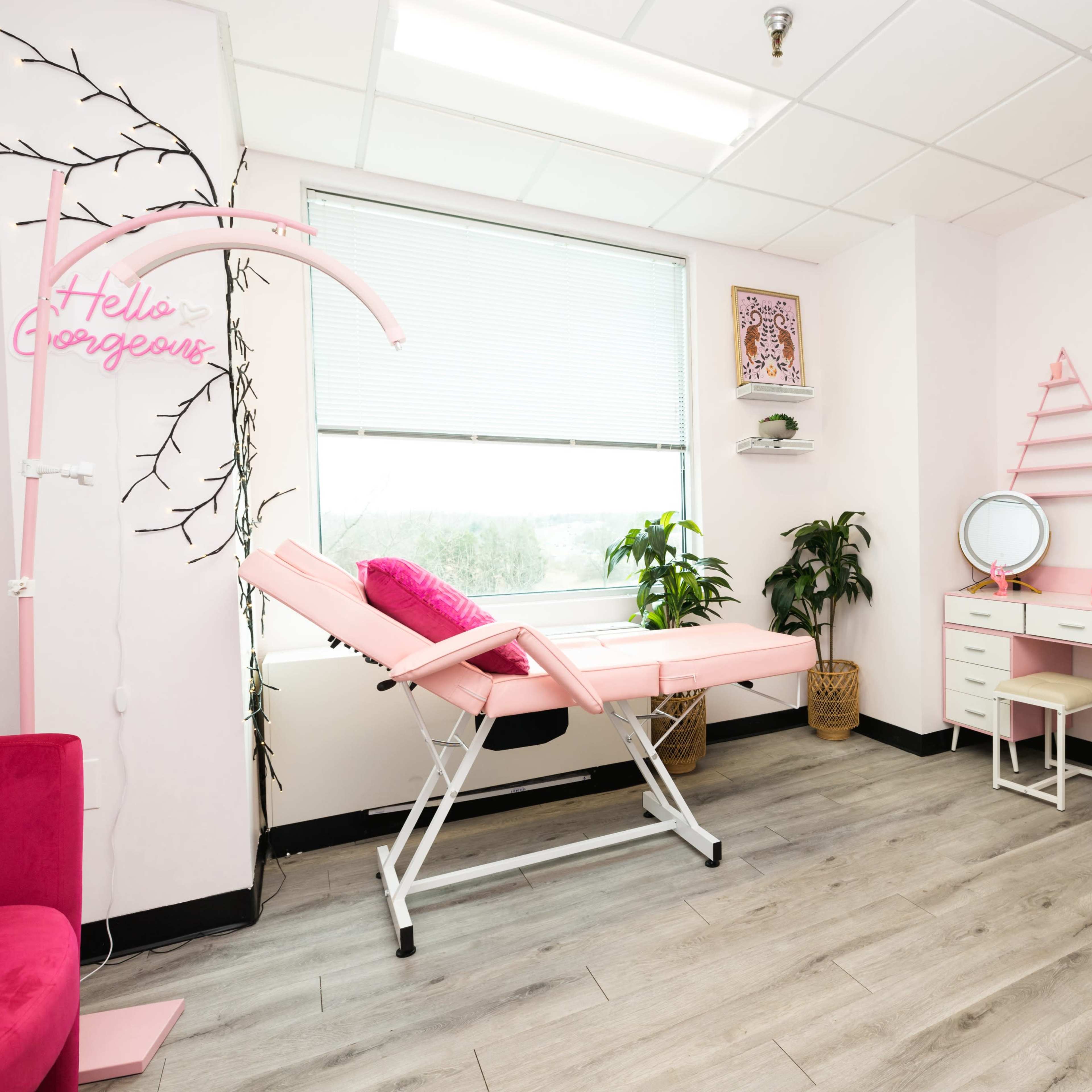 The room features a pink treatment bed, a small vanity desk, and a framed black-and-white portrait on the wall.