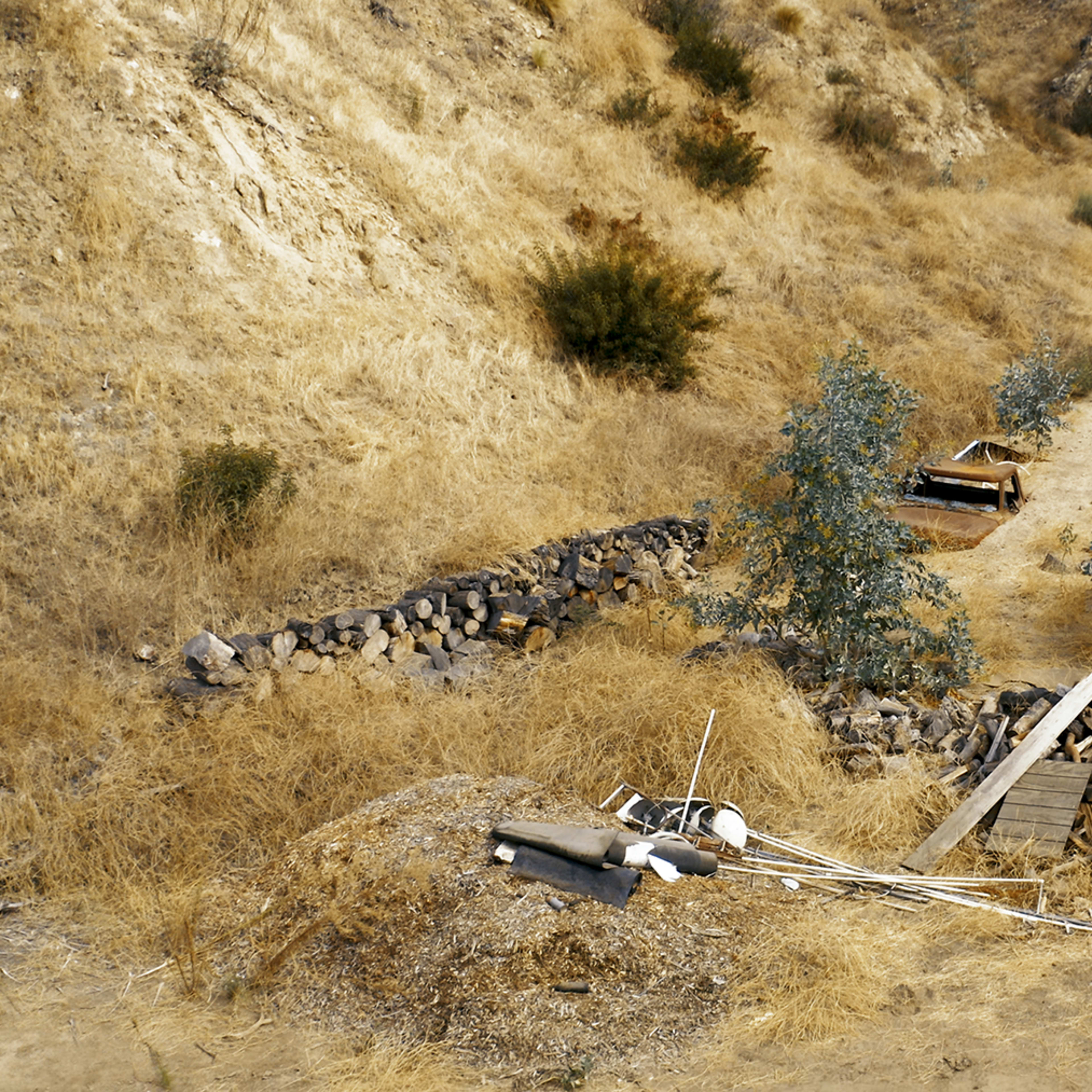 A dry, uneven landscape with scattered debris, including piles of wood and metal, surrounded by sparse vegetation and rocky ground.