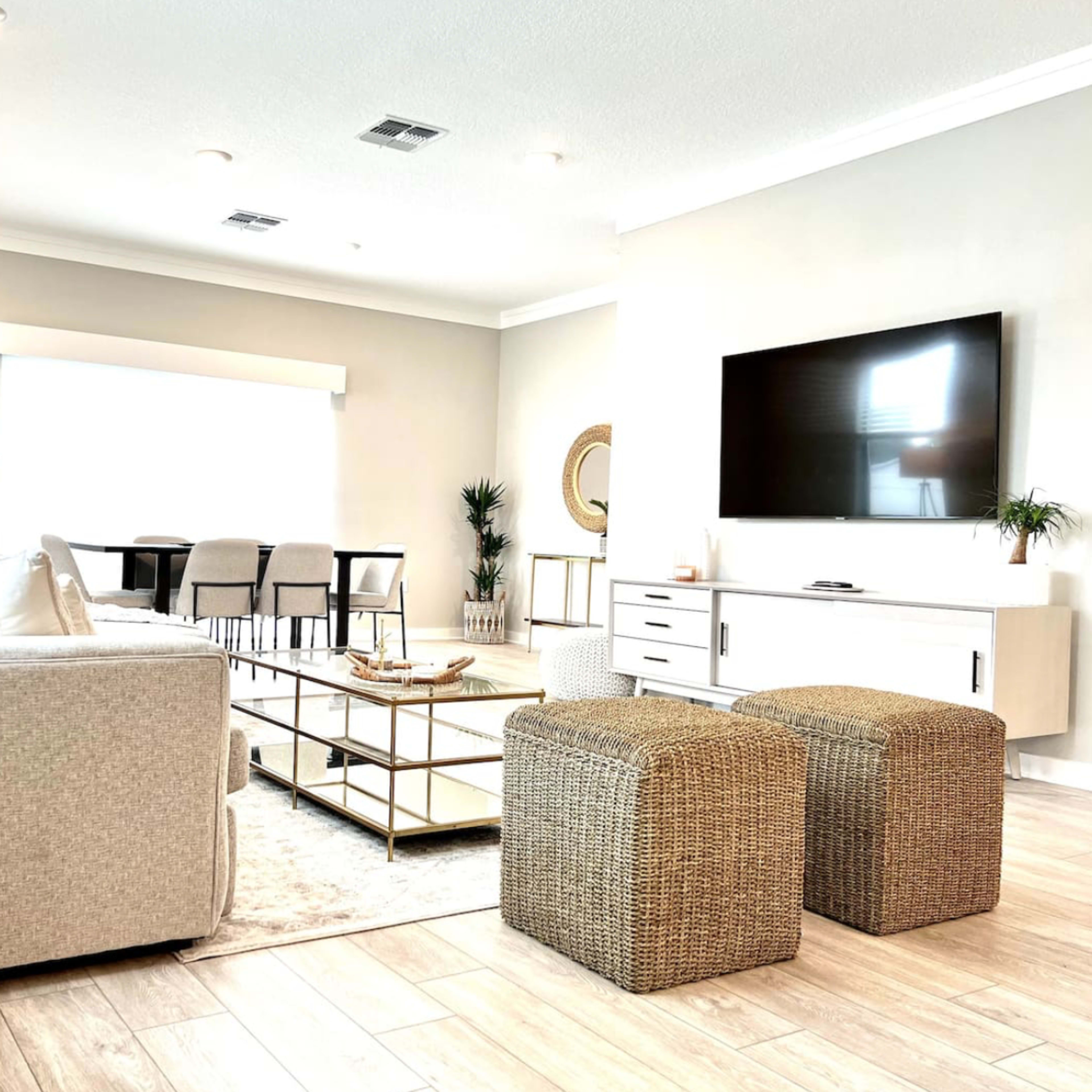 A modern living room features a light-colored sofa, a coffee table, two woven ottomans, and a large TV mounted on the wall.