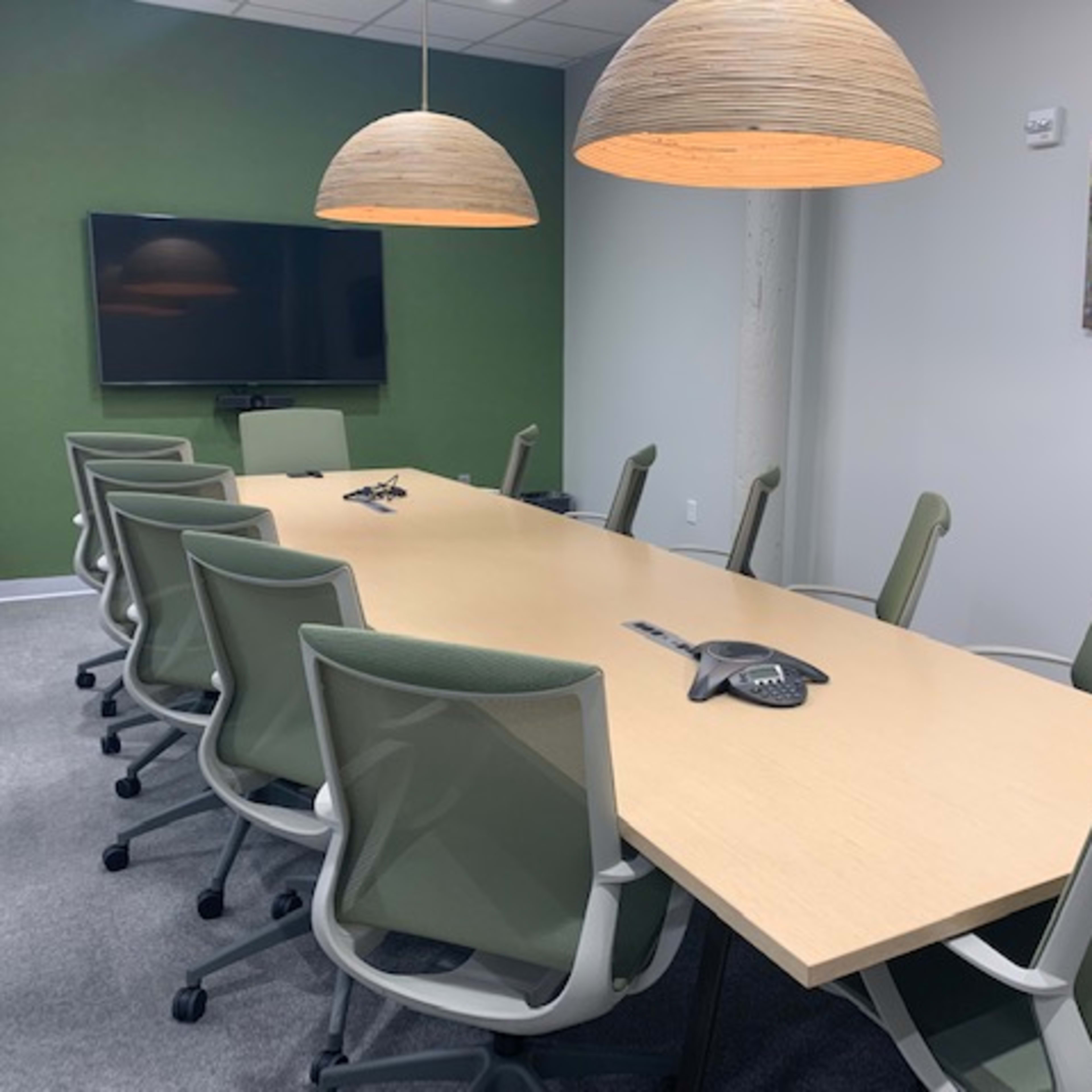 A long wooden conference table with eight green office chairs is positioned under two pendant lights in a modern meeting room adorned with a green accent wall.