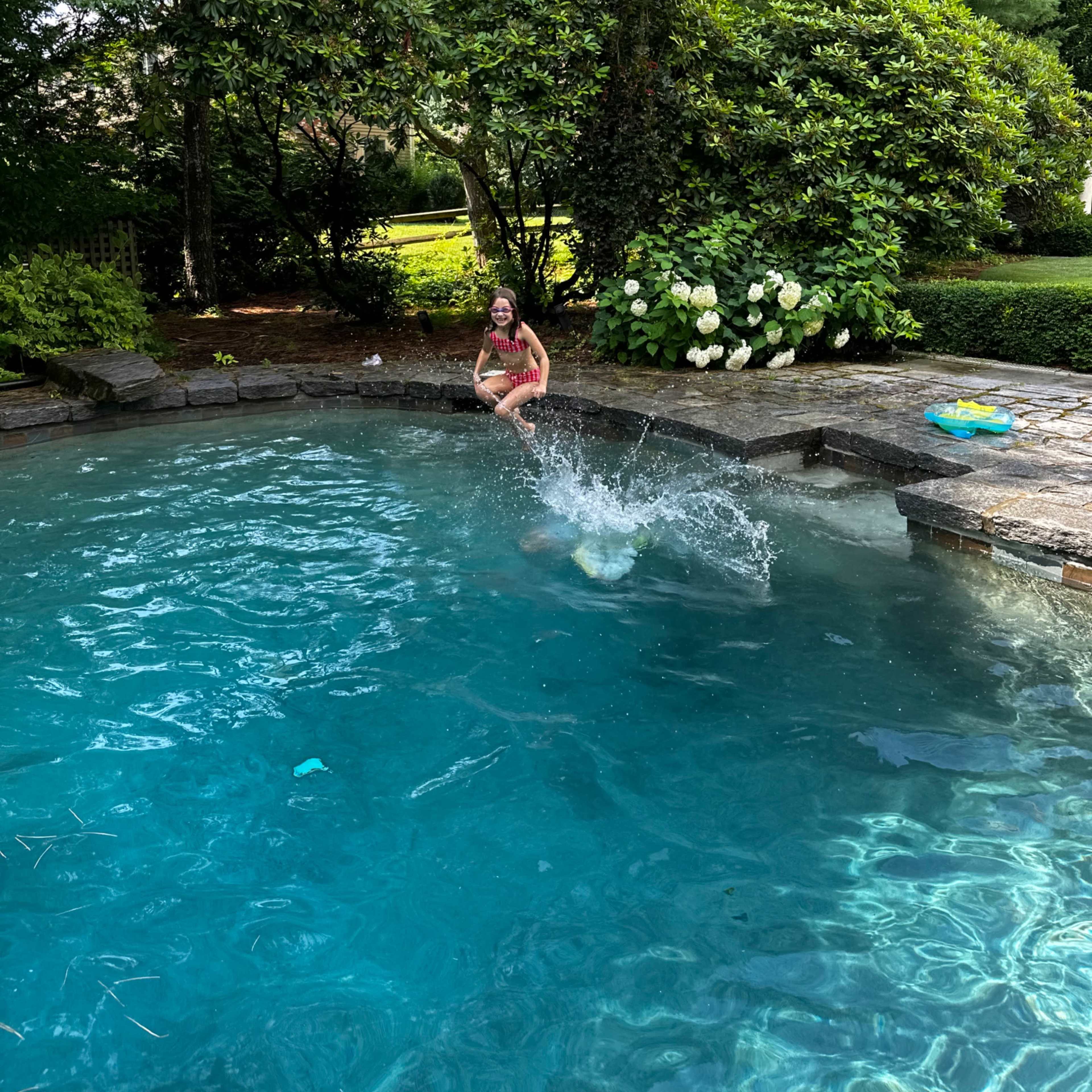 A child is sitting on the edge of a pool while another child splashes into the water.