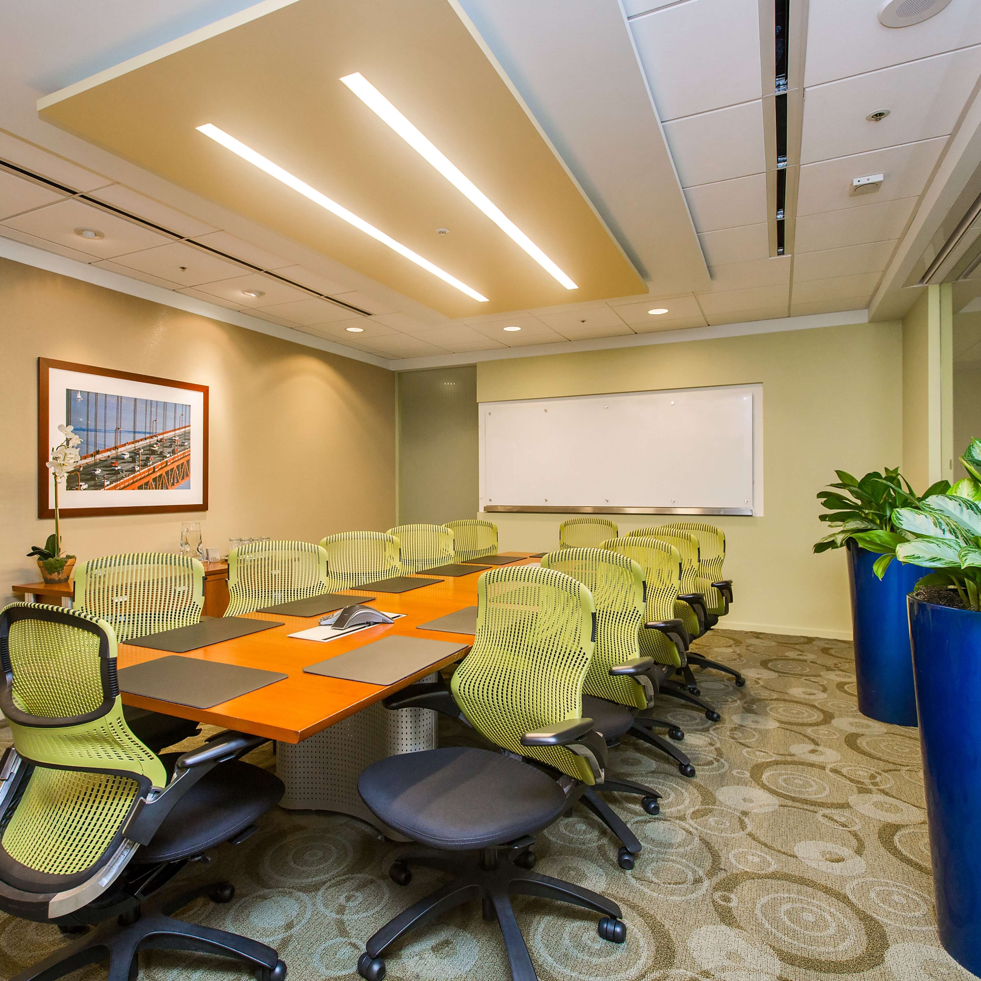 A modern conference room with a long table surrounded by ergonomic chairs, a whiteboard on one wall, and large potted plants in the corners.