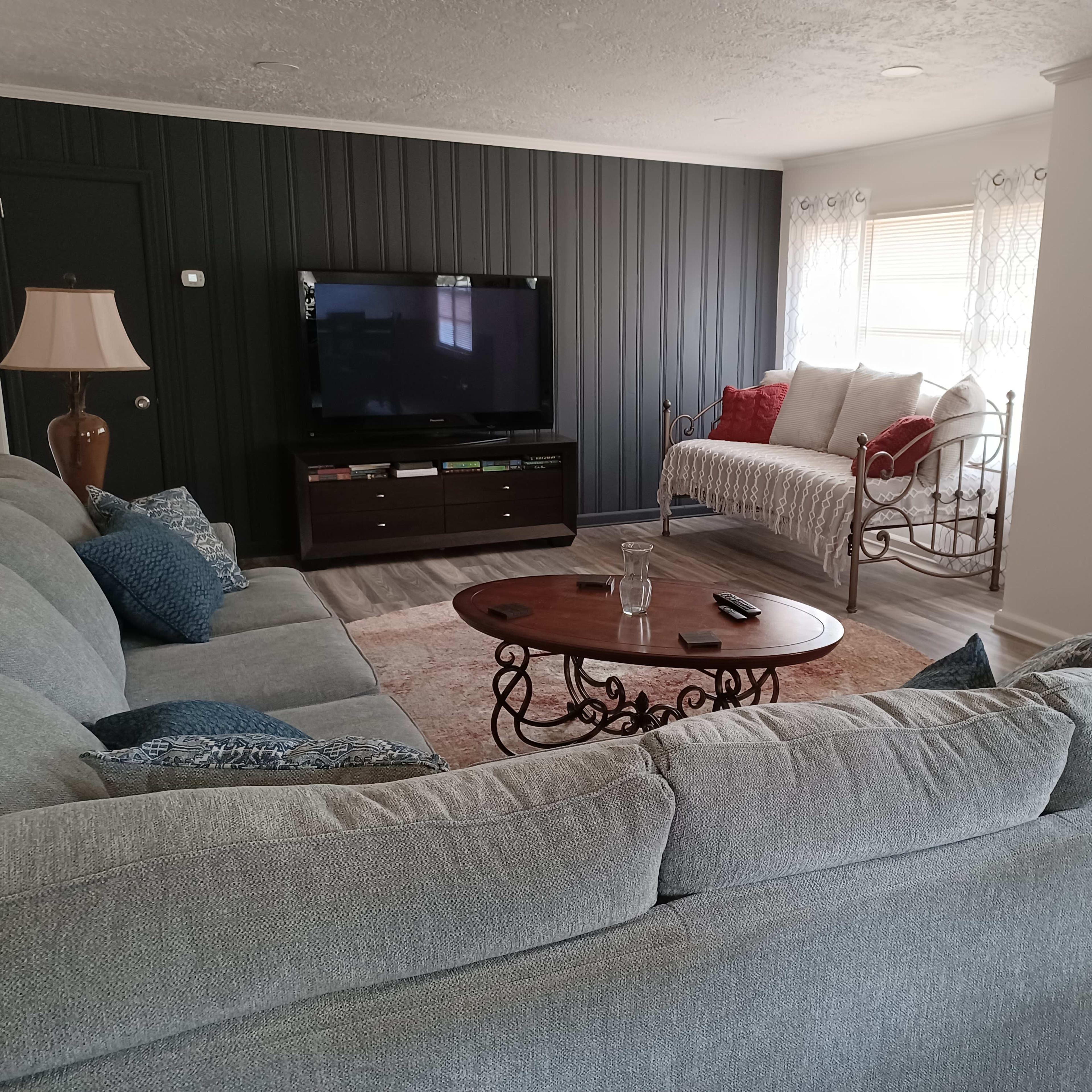 A living room features a gray sectional sofa, a coffee table, a television on a dark cabinet, and a daybed near a teal-colored door.