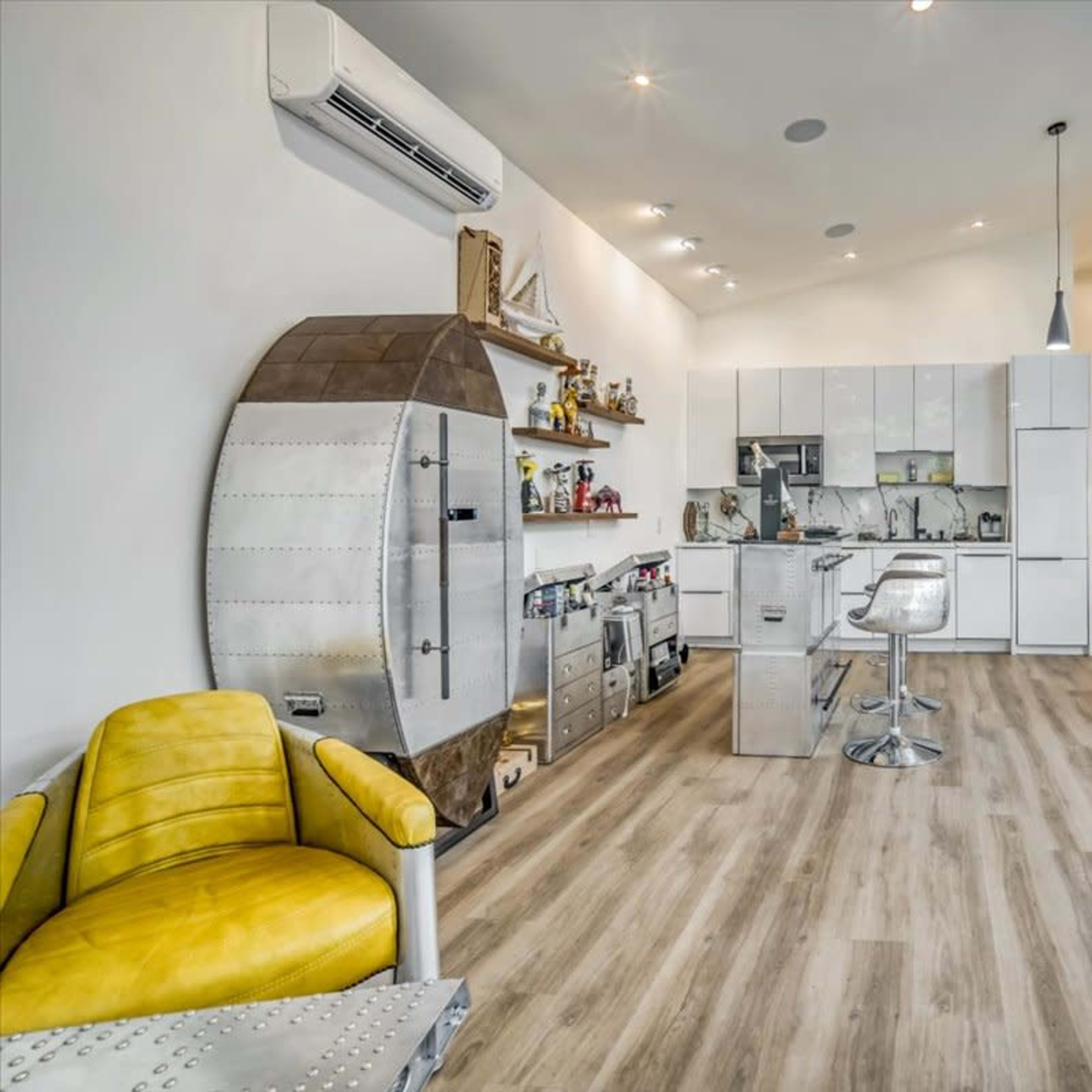 A modern room with light wood flooring, large windows, and a mix of furniture including a yellow chair and metallic accents.