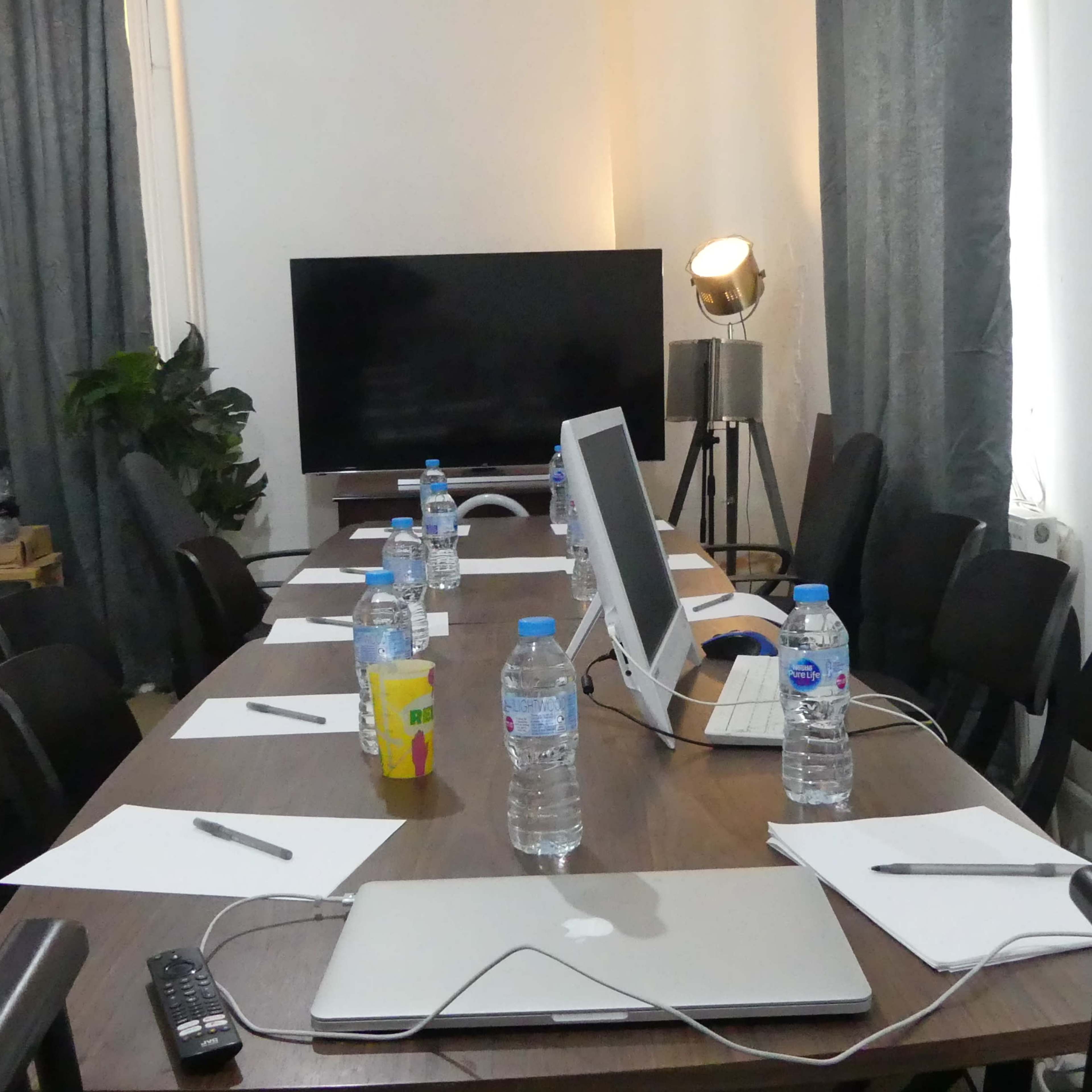 A conference room is set up with a long table, several chairs, bottles of water, notepads, and electronic devices, including a computer and a television.