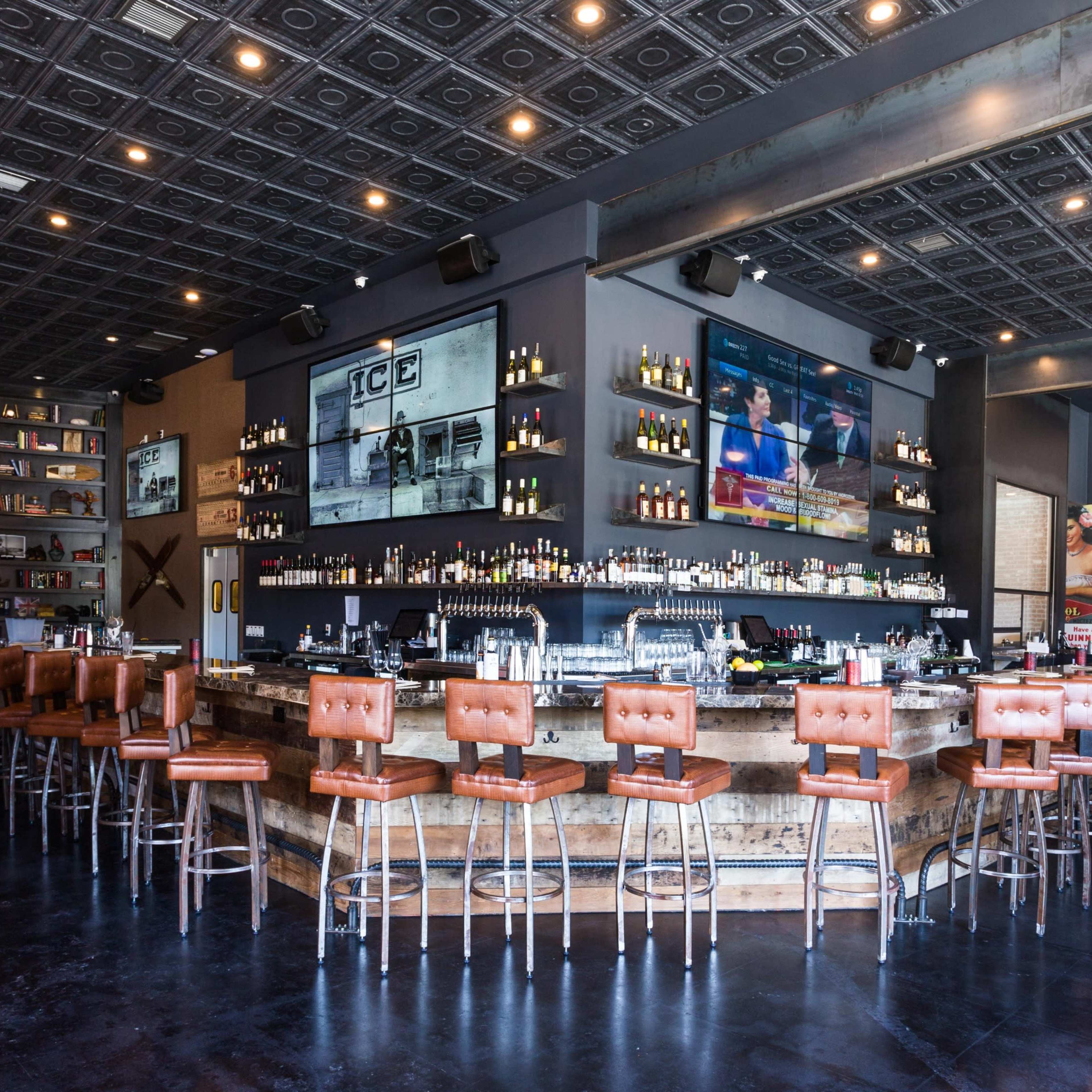 A well-lit restaurant bar features a long wooden counter with leather stools, surrounded by shelves filled with books and various bottles, complemented by large TVs mounted on the walls.