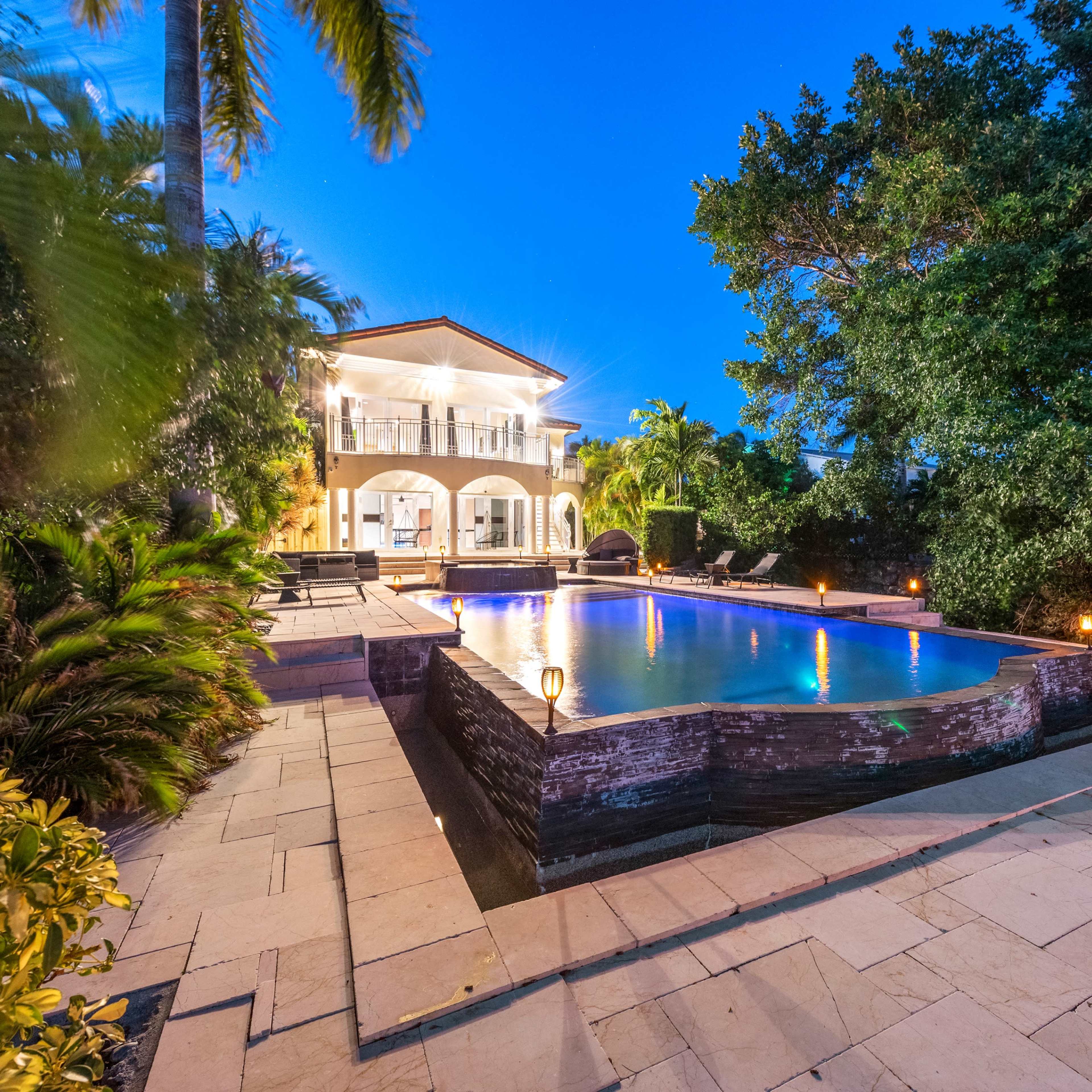 A backyard features a swimming pool illuminated by lights, surrounded by palm trees and a two-story house.