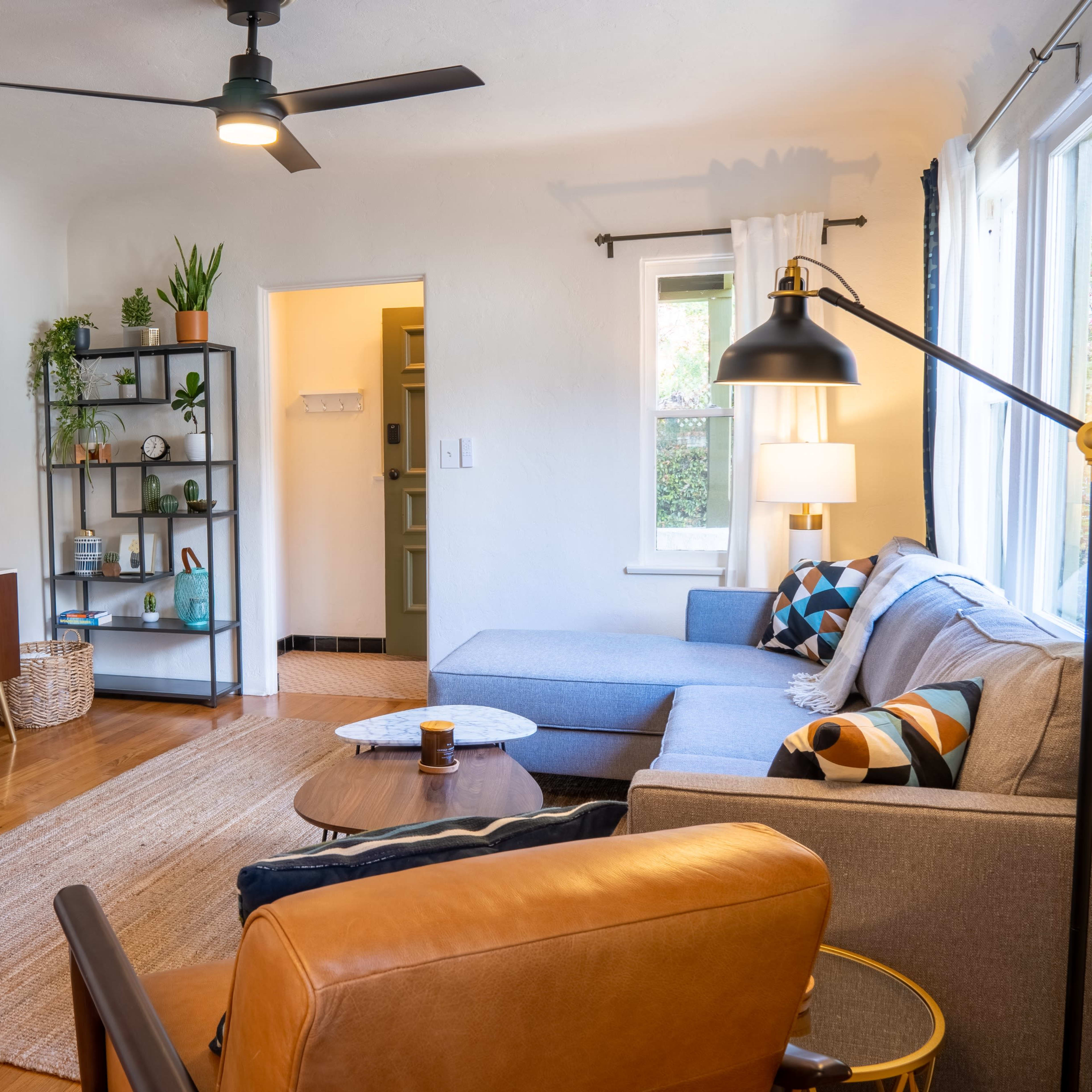 The living room features a sectional sofa, two armchairs, a coffee table, a television mounted on the wall, and a shelf adorned with plants and decorative items.