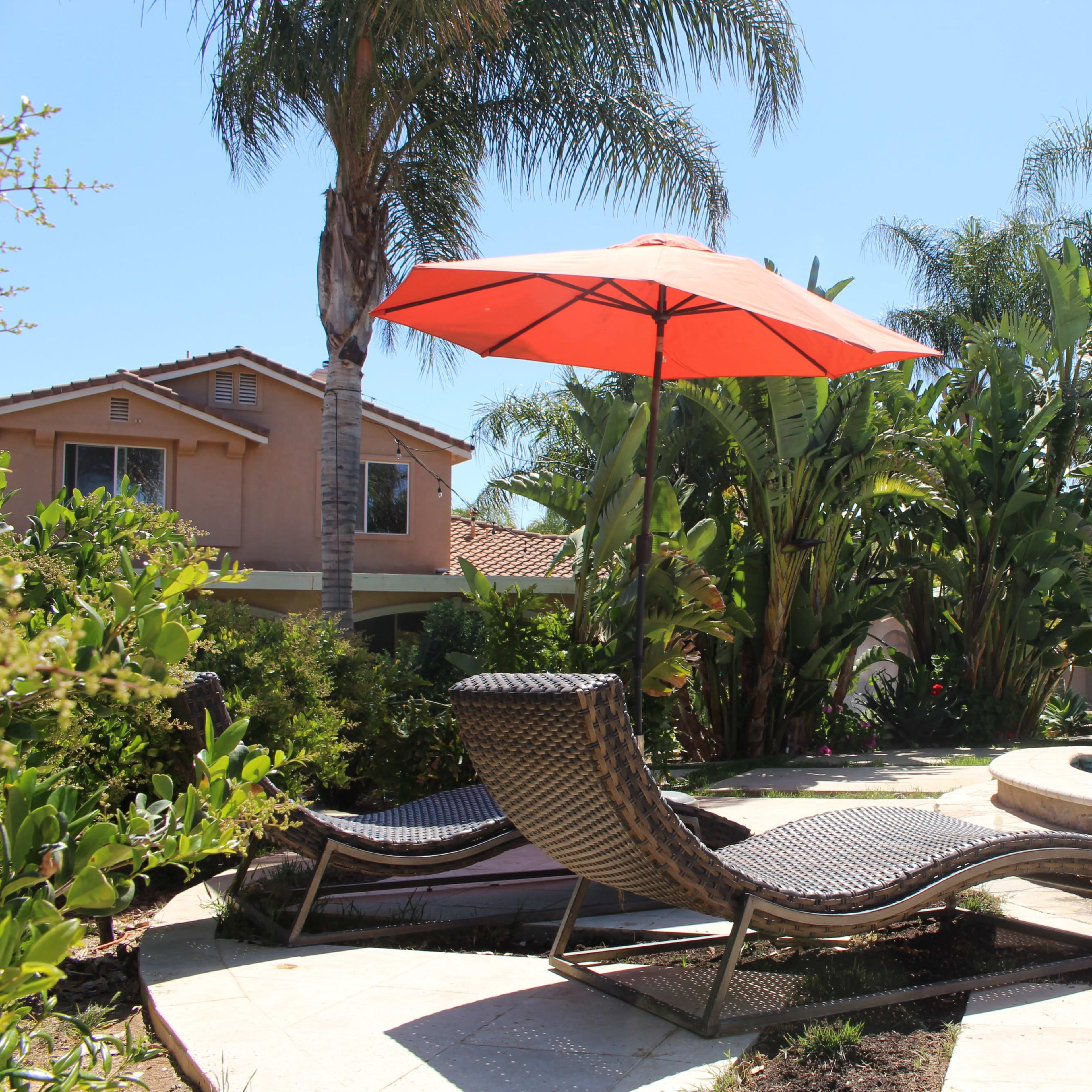 A poolside area features two lounge chairs beneath a large orange umbrella, surrounded by tropical plants and a residential house in the background.