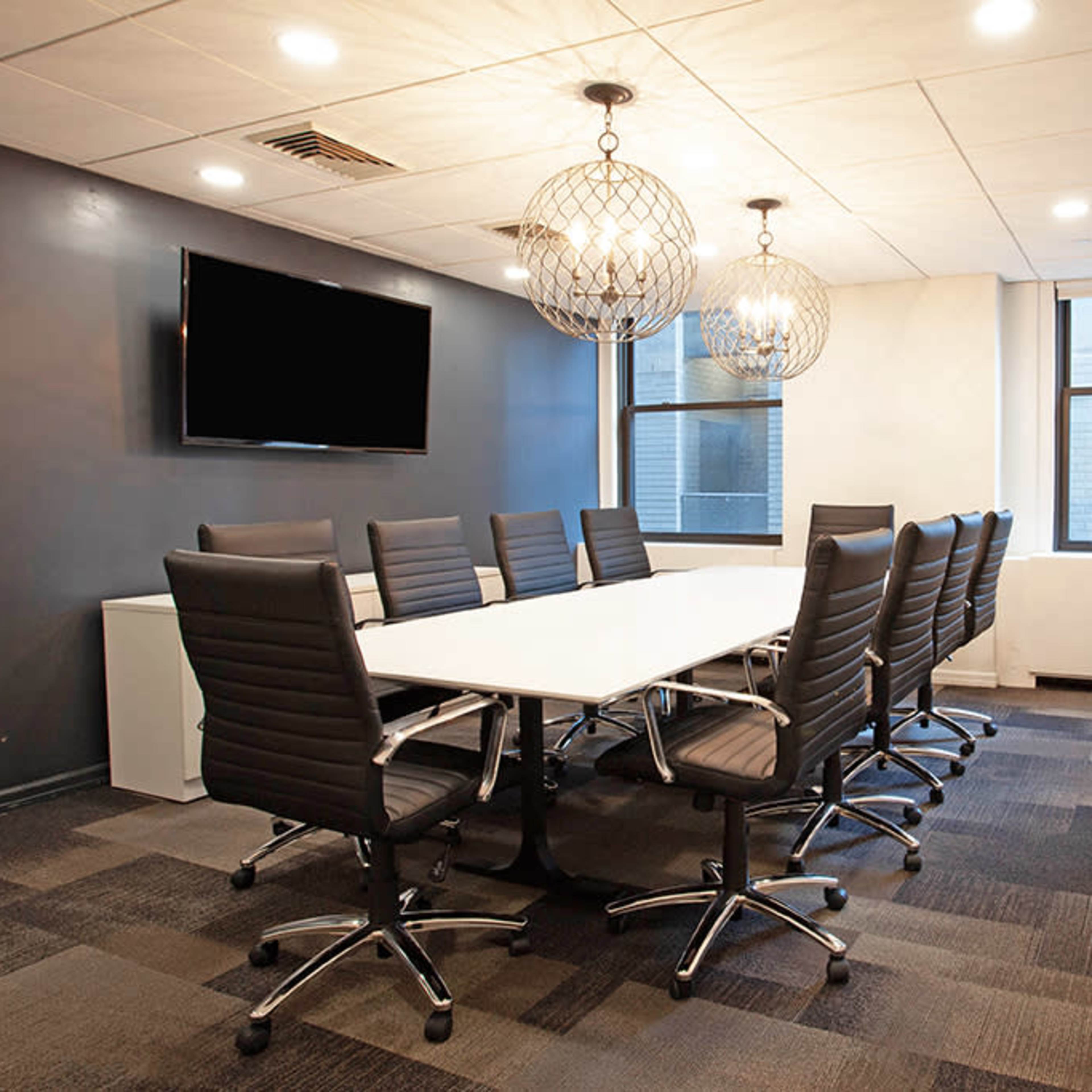 A modern conference room features a long white table surrounded by black swivel chairs, with pendant lighting and a wall-mounted TV.