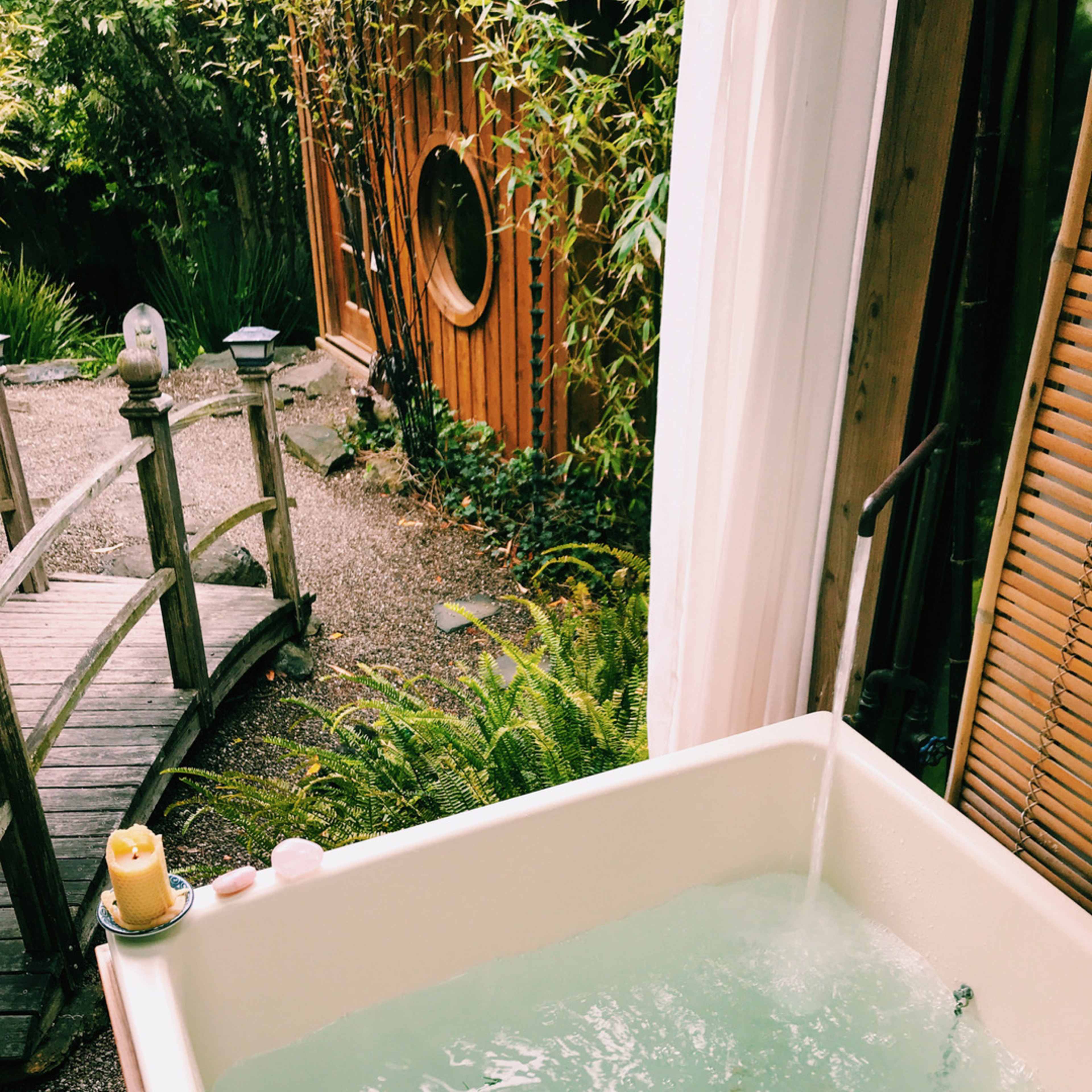 A wooden bathtub filled with water and floating petals is placed next to a garden with a small bridge and bamboo greenery.