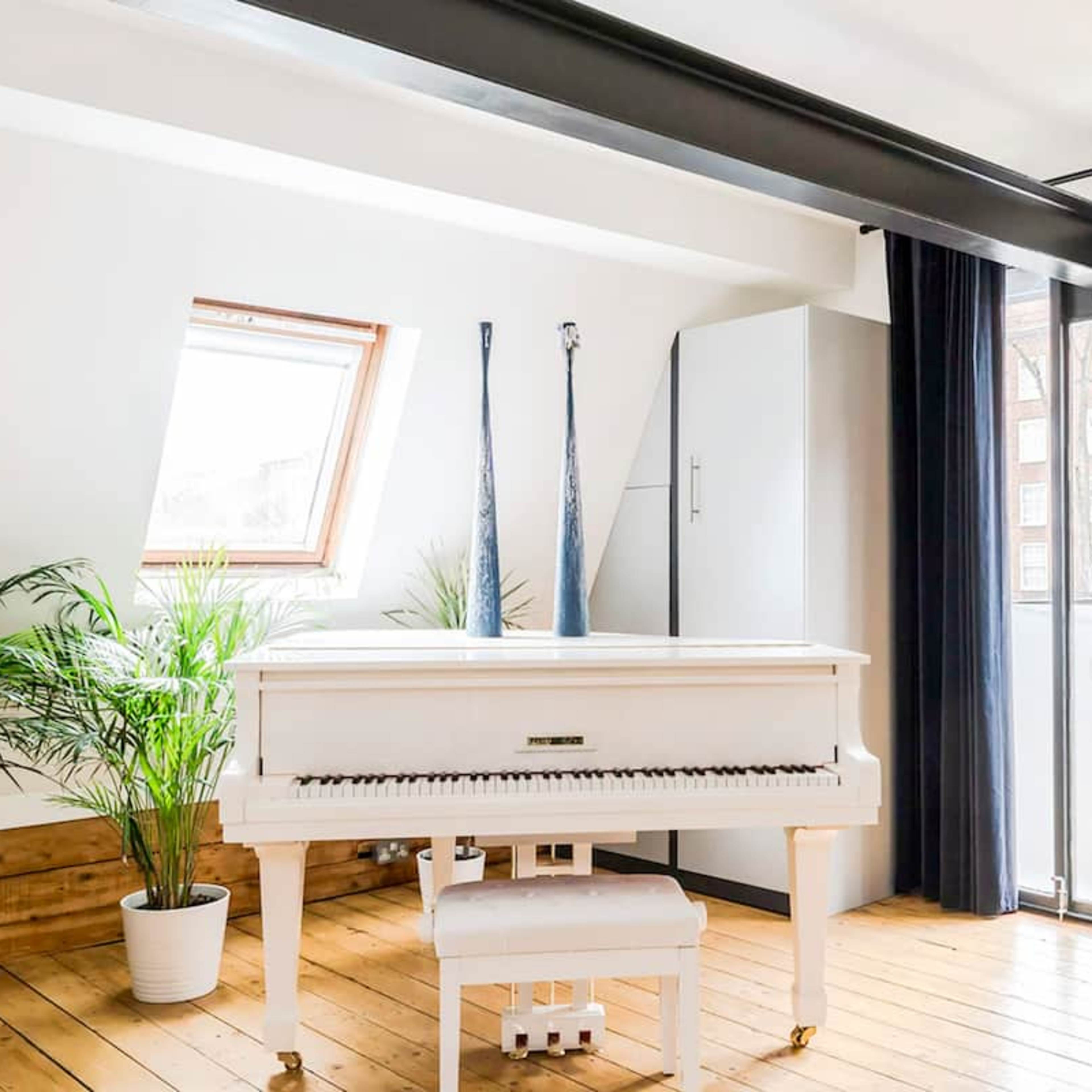 A white piano is positioned in a bright, airy room with wooden flooring, potted plants, and large windows.