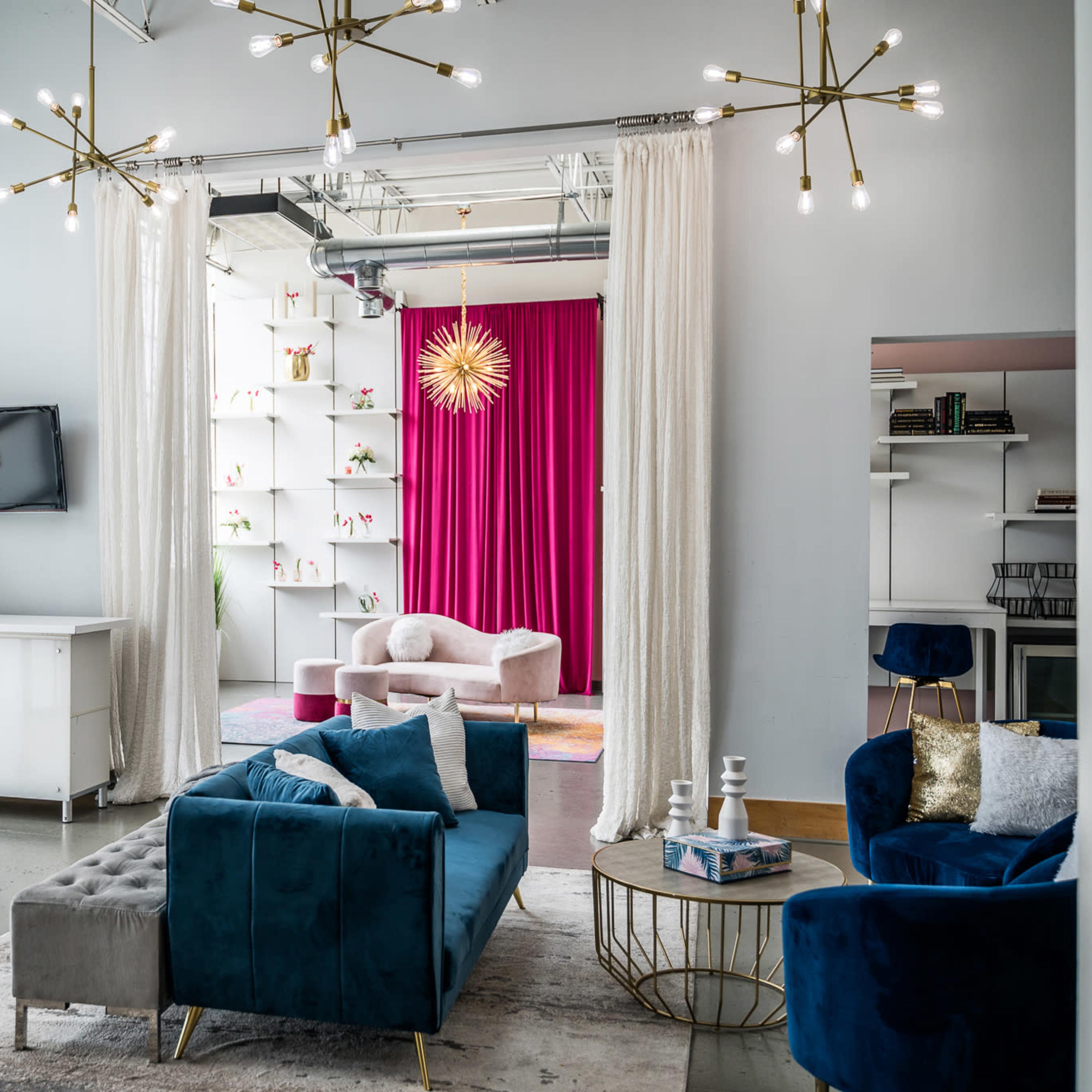 A modern interior space features a living area with a blue velvet sofa, a coffee table, and a white cabinet, with a backdrop of pink curtains and decorative shelves.