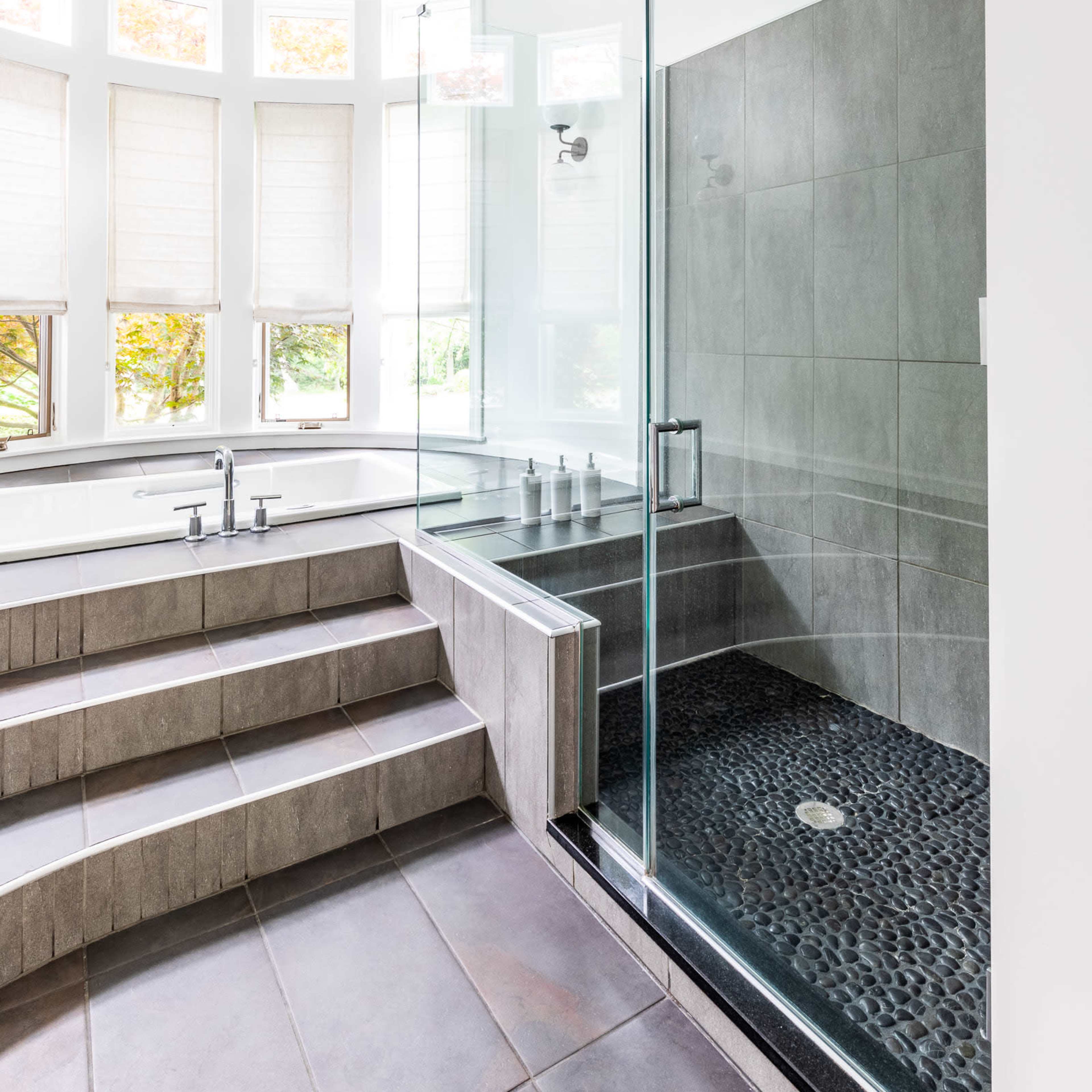 A modern bathroom featuring a glass-enclosed shower with pebble flooring and a deep soaking tub next to a large window with multiple panels.