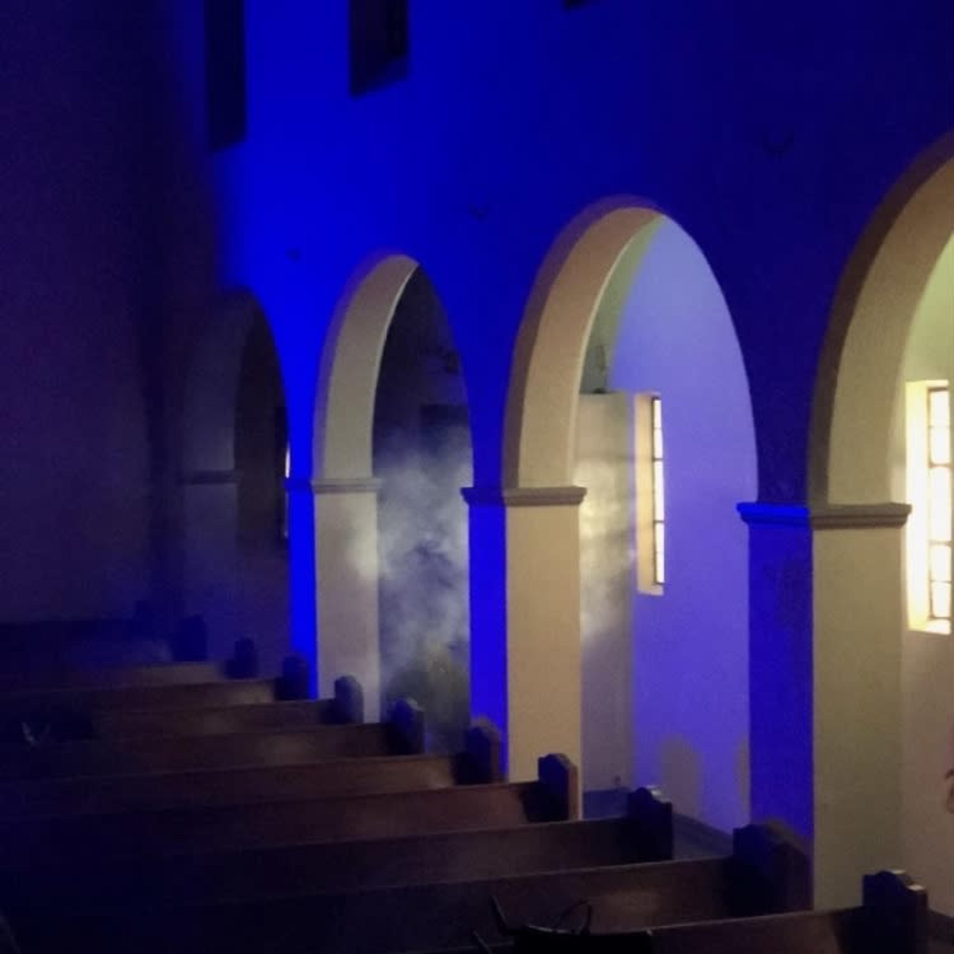 A dimly lit church interior features arched doorways and pews, with blue lighting creating a misty atmosphere.