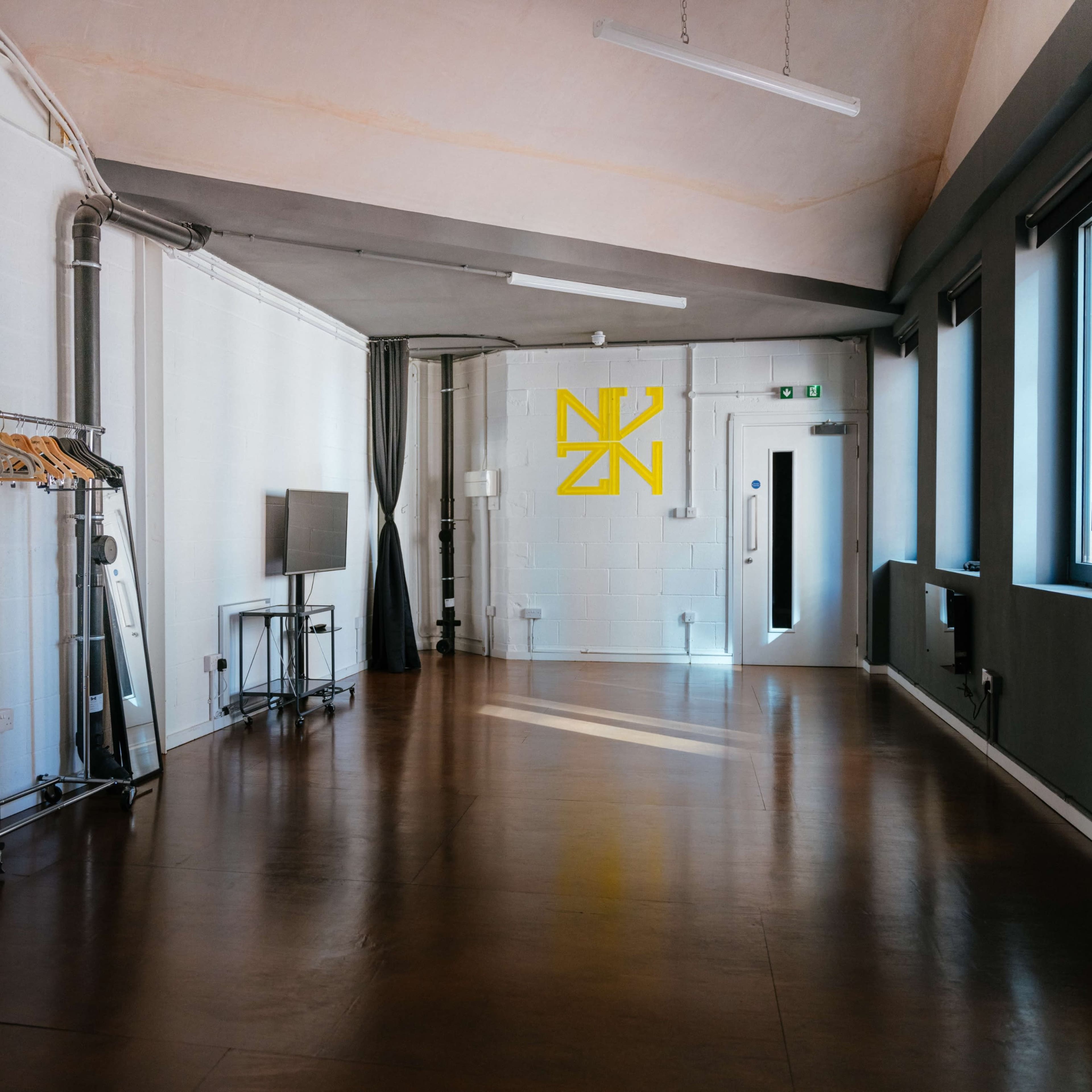 An empty room with a polished floor, a clothing rack on wheels, a television on a stand, and a yellow logo on a white wall.